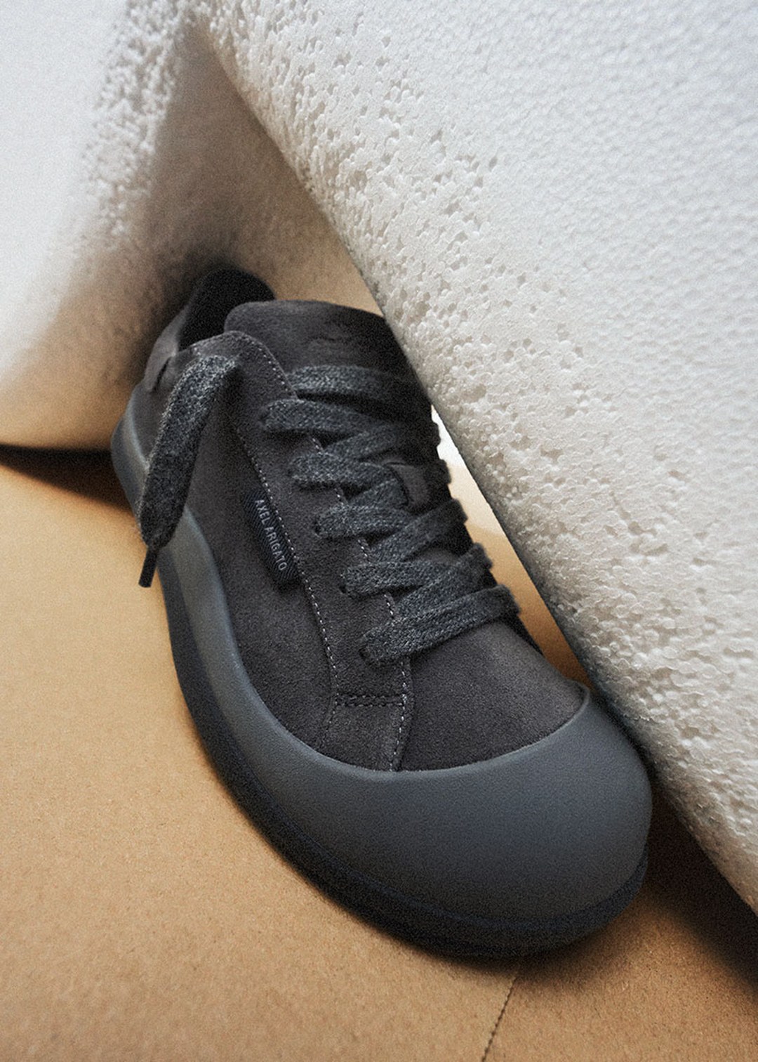A dark gray suede sneaker with black laces, placed against a textured white and brown background.
