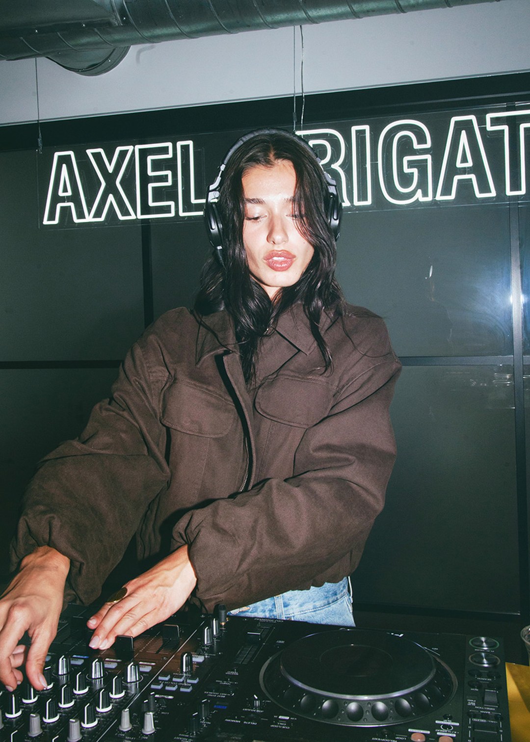 A DJ wearing headphones mixes on a soundboard, set against a backdrop with illuminated text.