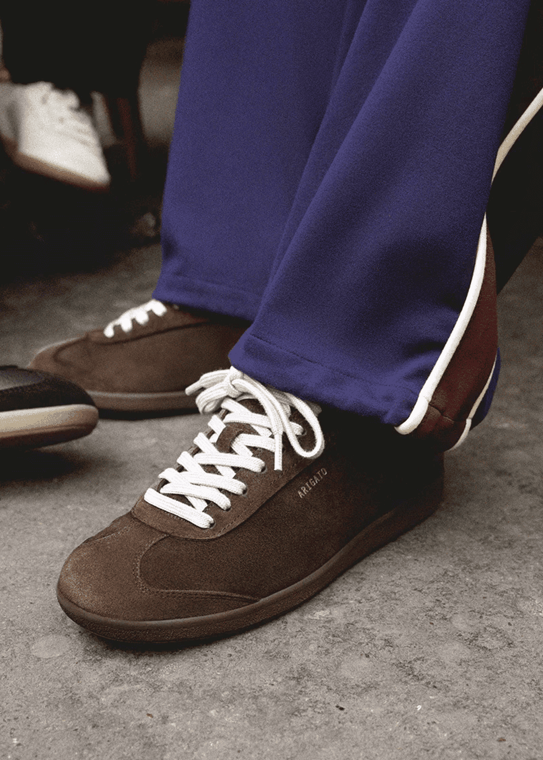 Brown suede sneakers with white laces are worn with blue pants having white trim, standing on a concrete floor.