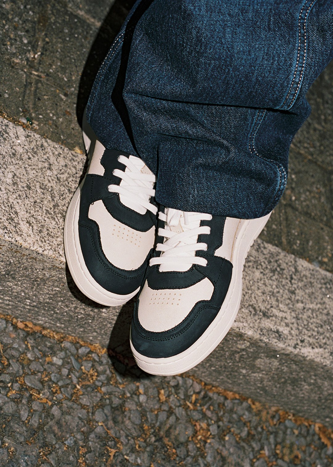 Person wearing white and black sneakers with dark jeans, standing on a textured concrete surface with pebbles and a curb.