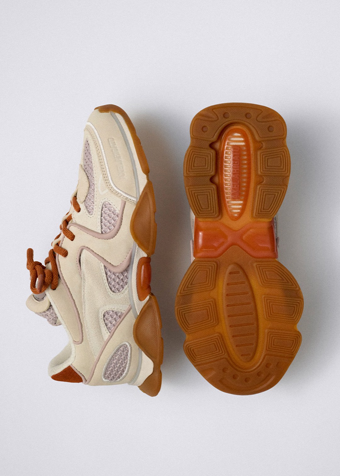 A light beige athletic shoe shown from the side and bottom, featuring orange laces and a textured gum sole against a plain background.