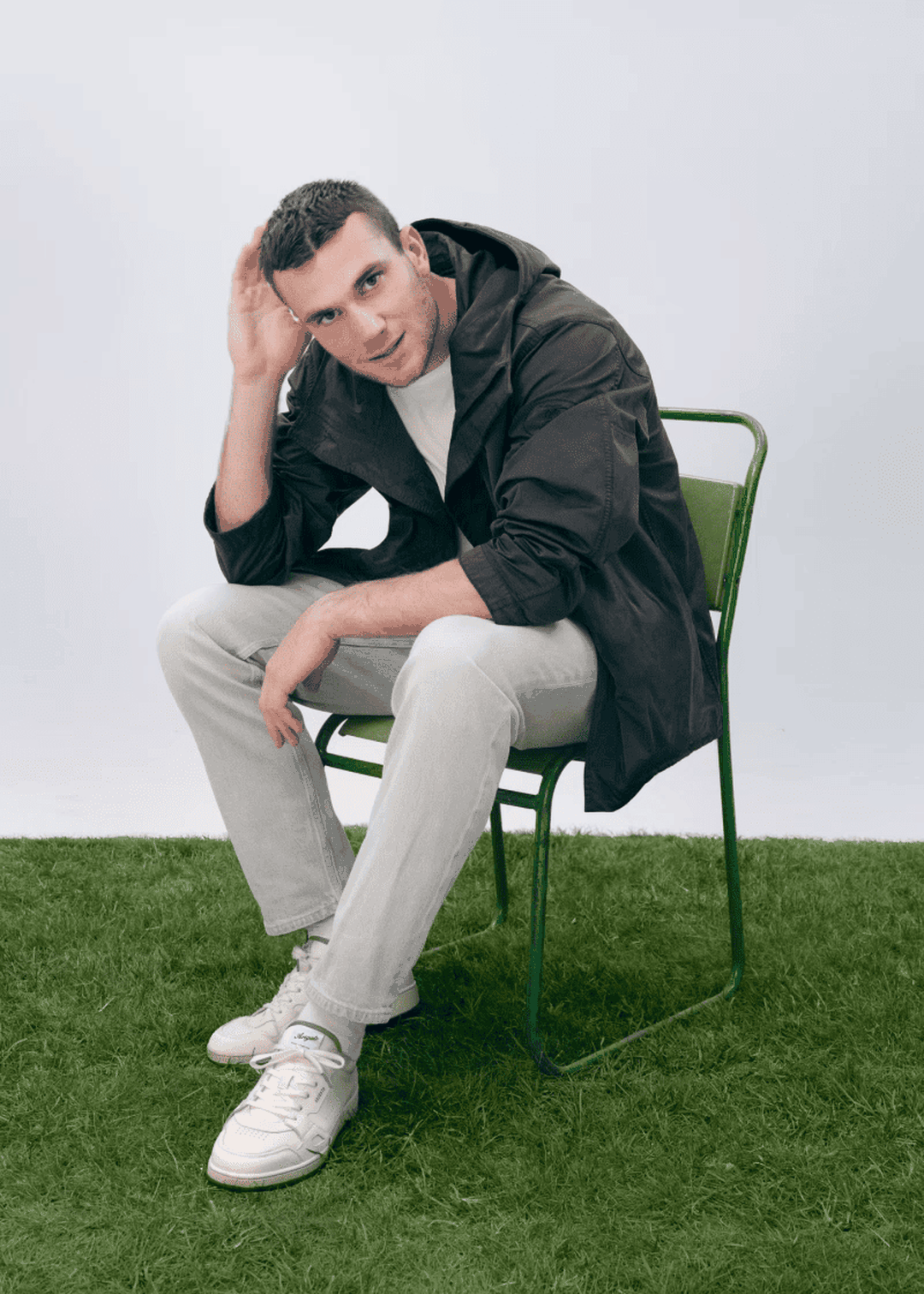 Man in casual attire sits on a green chair on grass, wearing a dark jacket, light pants, and sneakers, with a thoughtful expression.