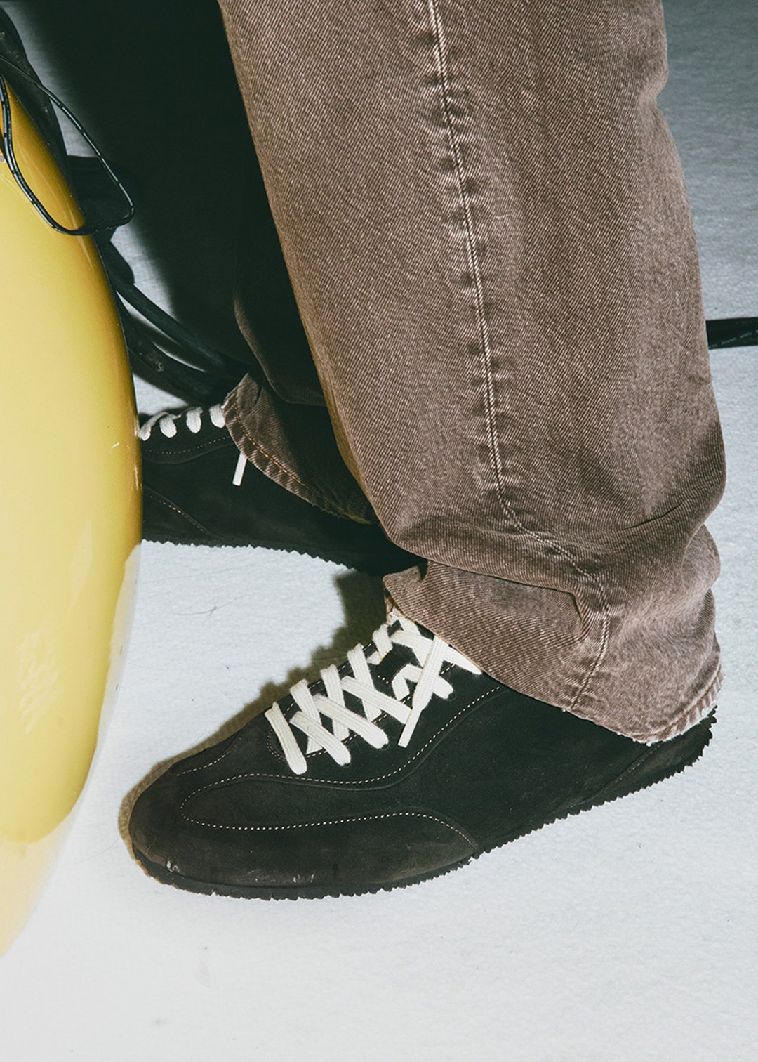 Person wearing brown pants and black sneakers with white laces stands next to a yellow object on a light-colored floor.