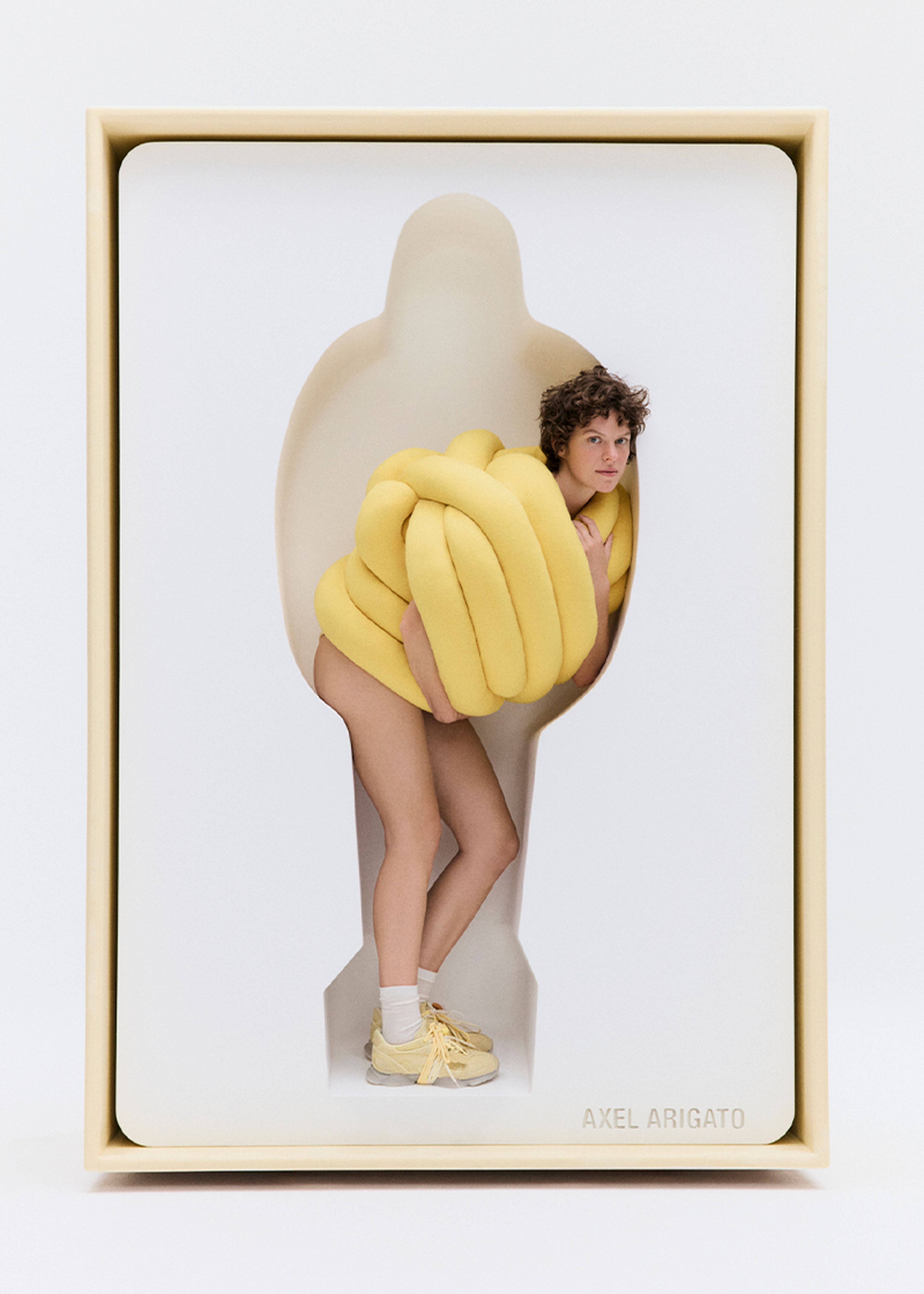 Person wearing a yellow, padded outfit stands in a silhouette-shaped cutout against a white background, with "AXEL ARIGATO" text below.