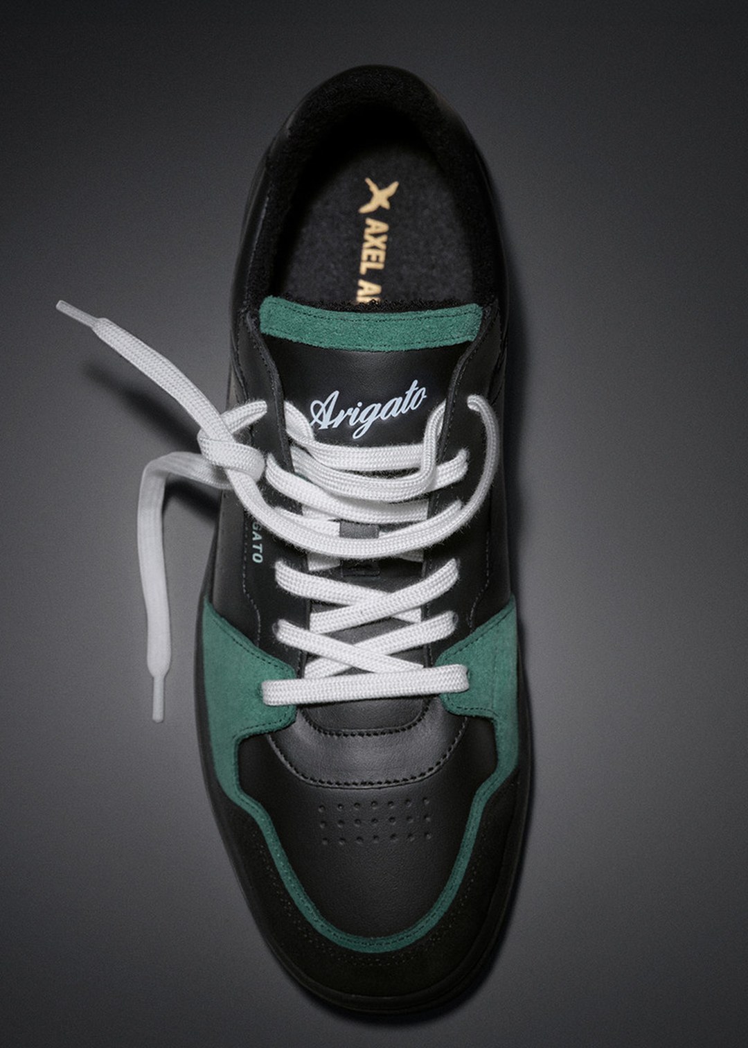 Black and green sneaker with white laces, featuring "Arigato" text on the upper, photographed from above on a dark surface.