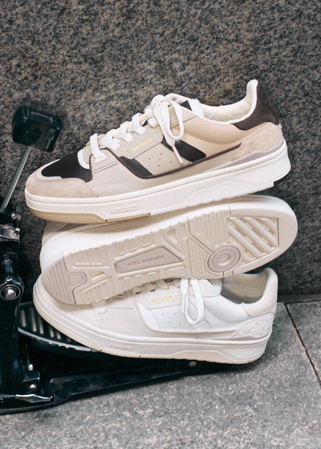 Two stacked sneakers on a metal pedal: one with brown and beige accents, the other in white. Both have textured soles and visible branding.