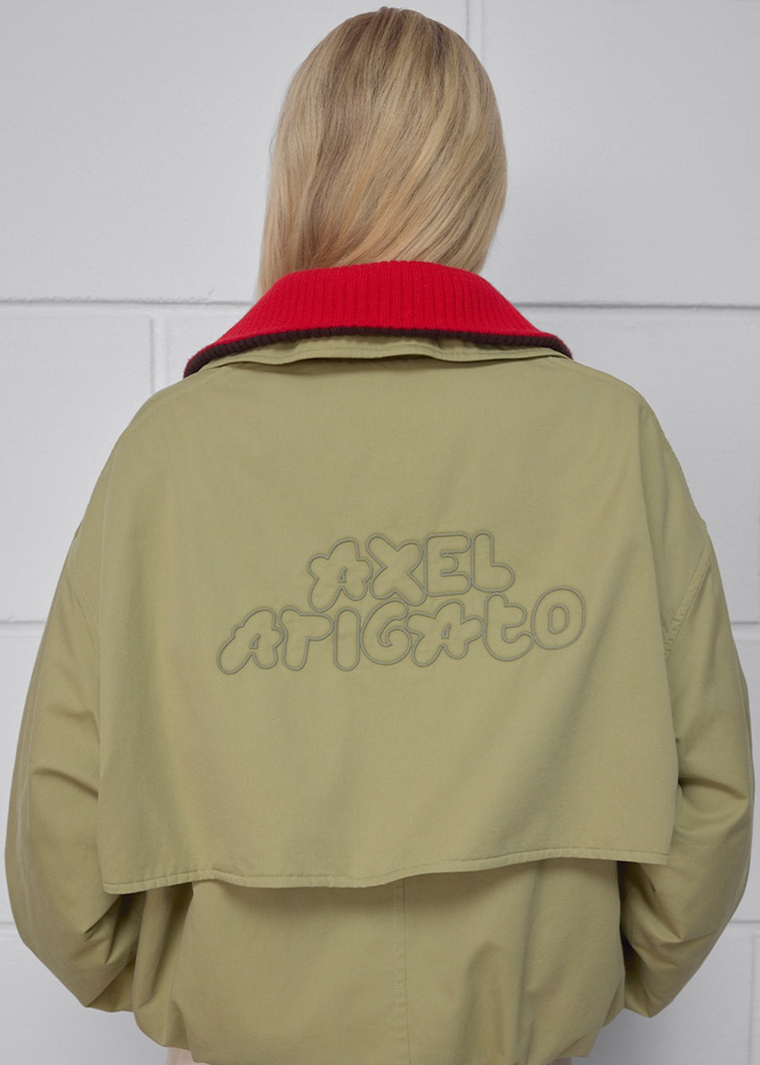 Person with long blonde hair wearing a beige jacket with "Axel Arigato" written on the back, red collar visible.