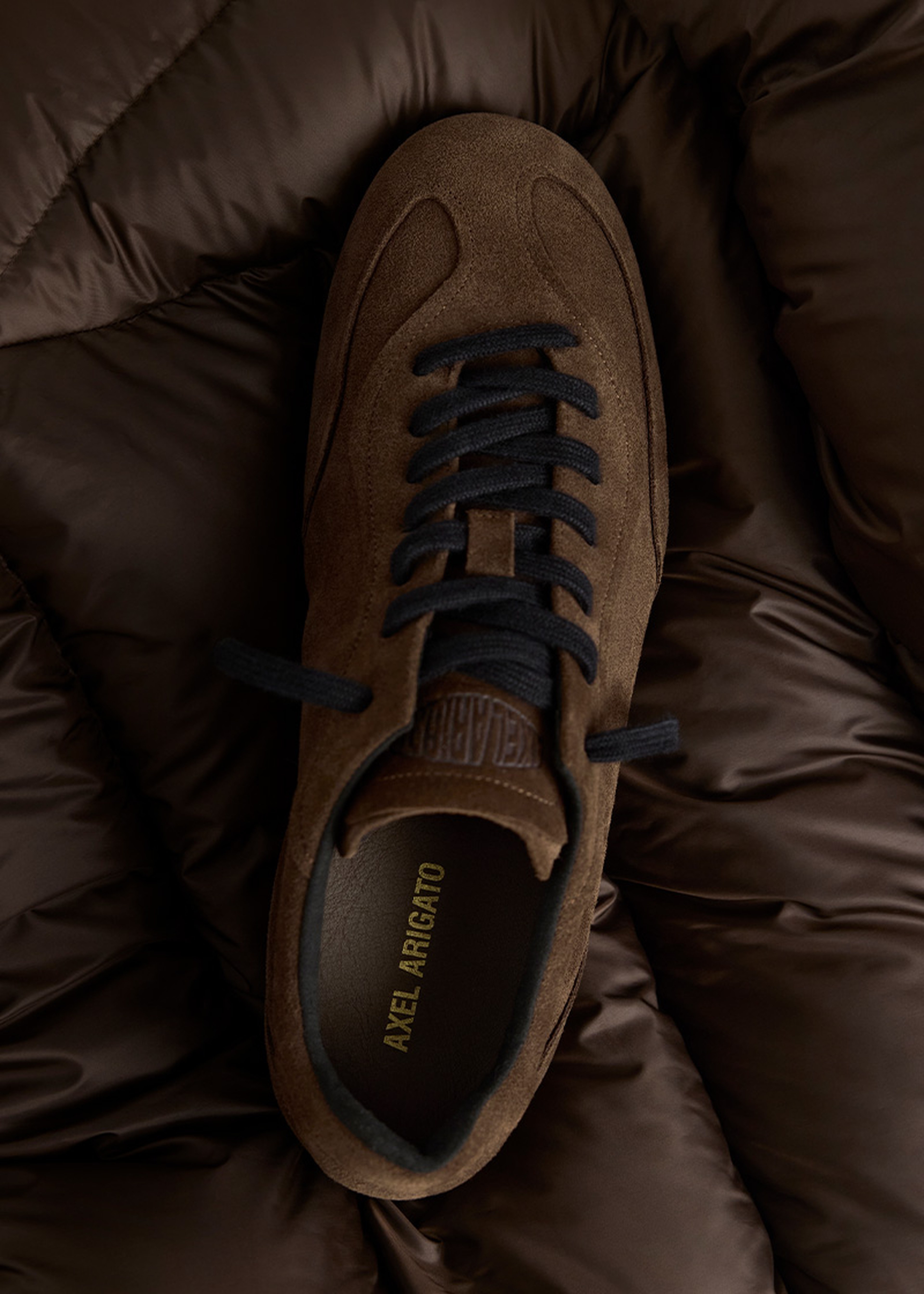 Brown suede sneaker with black laces on a dark, quilted background. "Axel Arigato" is visible inside the shoe.