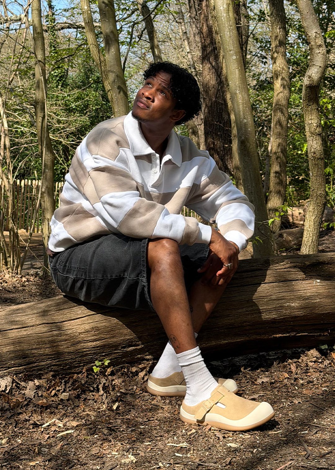 Person in a striped sweater and shorts sits on a log in a forested area, looking up thoughtfully at the sky.