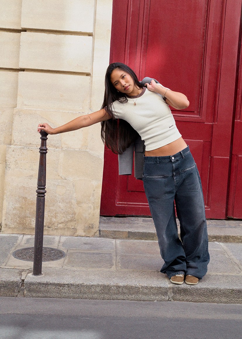 Woman in a crop top and wide-leg jeans leans playfully on a post against a red door backdrop.