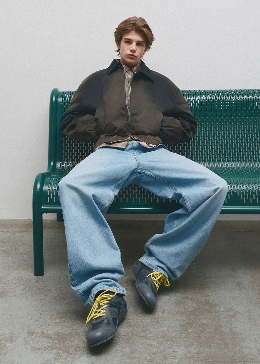 Person sitting on a green bench wearing a dark jacket, loose blue jeans, and sneakers with yellow laces.