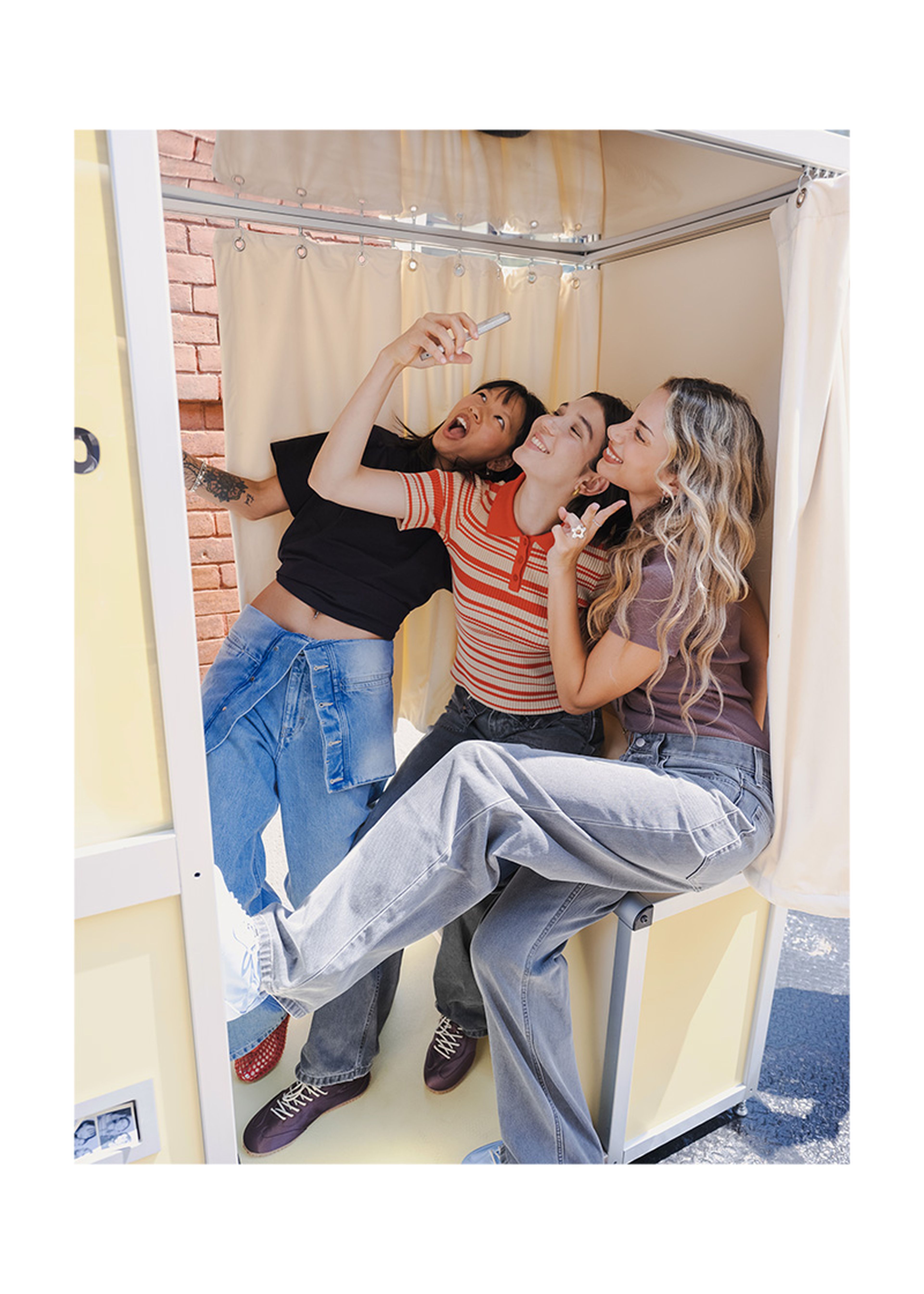 Three friends pose playfully inside a photo booth, taking a selfie. They are smiling and dressed casually, with one person lifting their foot.