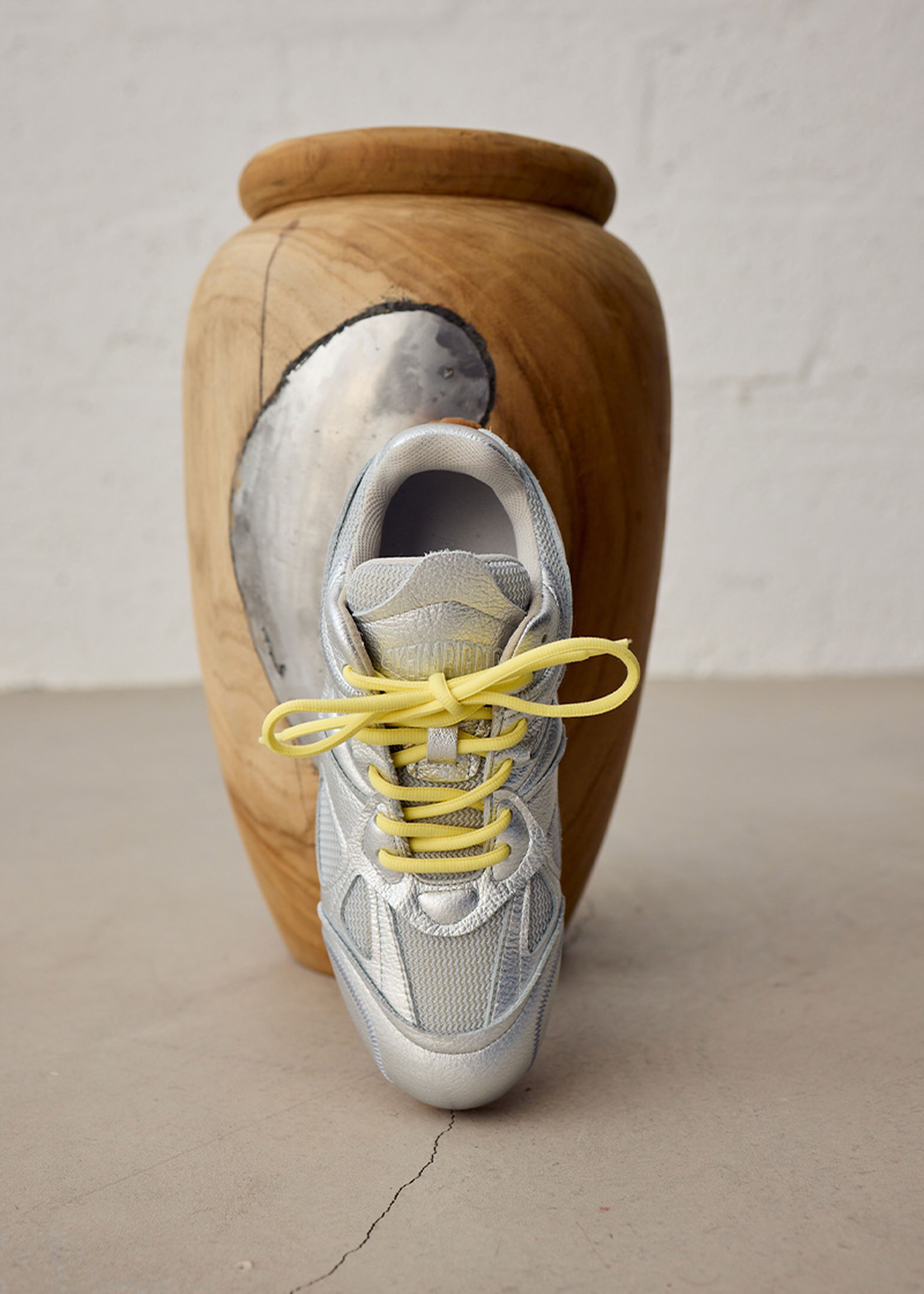 A single gray sneaker with yellow laces placed in front of a large, wooden vase on a concrete floor.