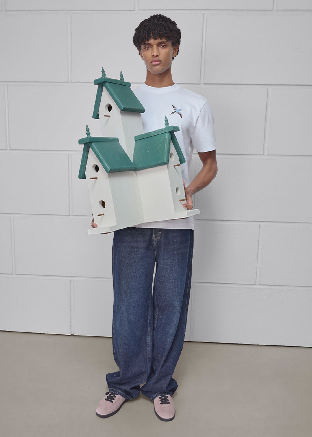 A person holding a large, multi-tiered birdhouse with green roofs, wearing a white t-shirt, blue jeans, and pink sneakers, standing indoors.