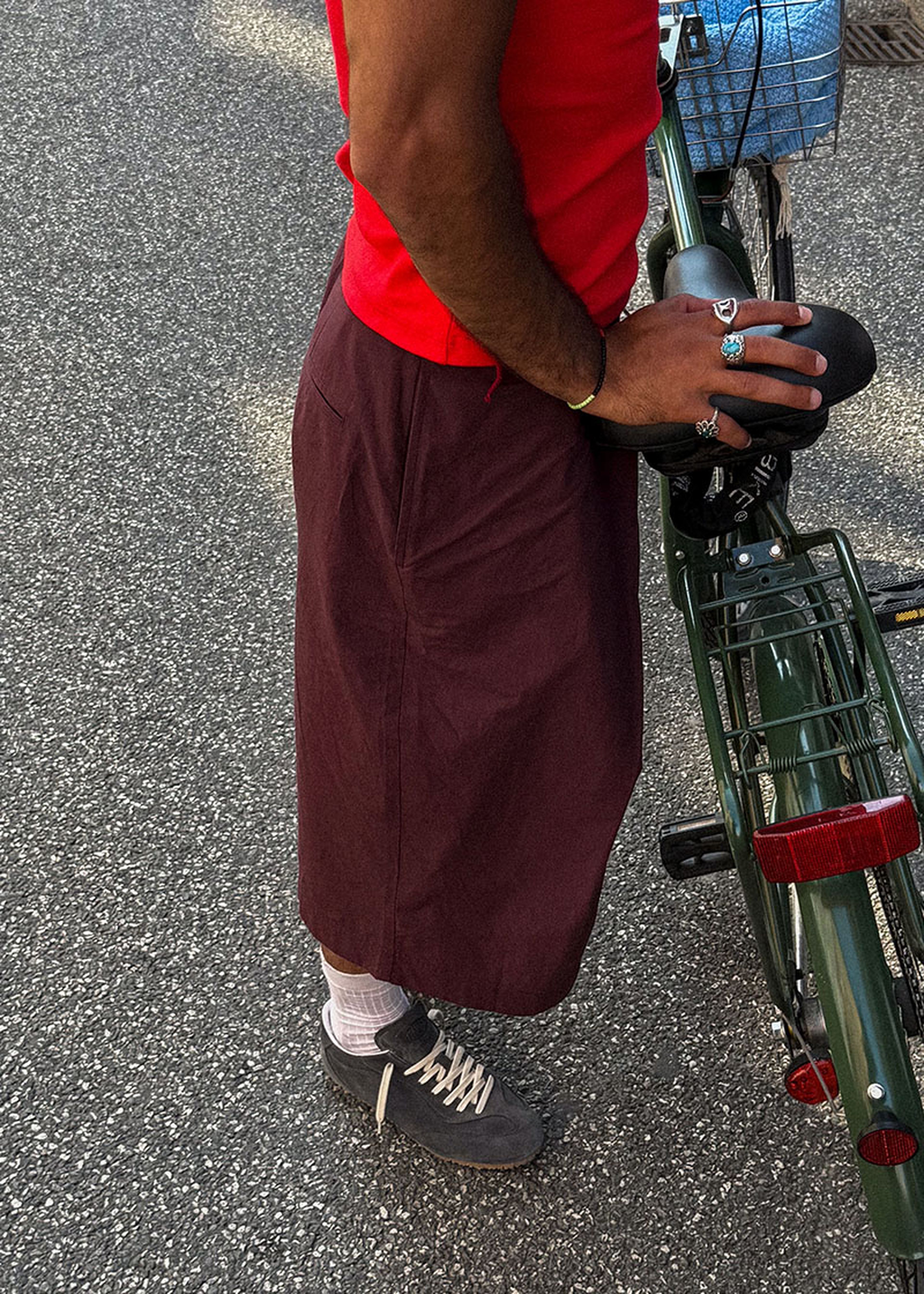 Person in a red shirt and maroon skirt stands beside a green bicycle on a paved surface, holding a helmet.