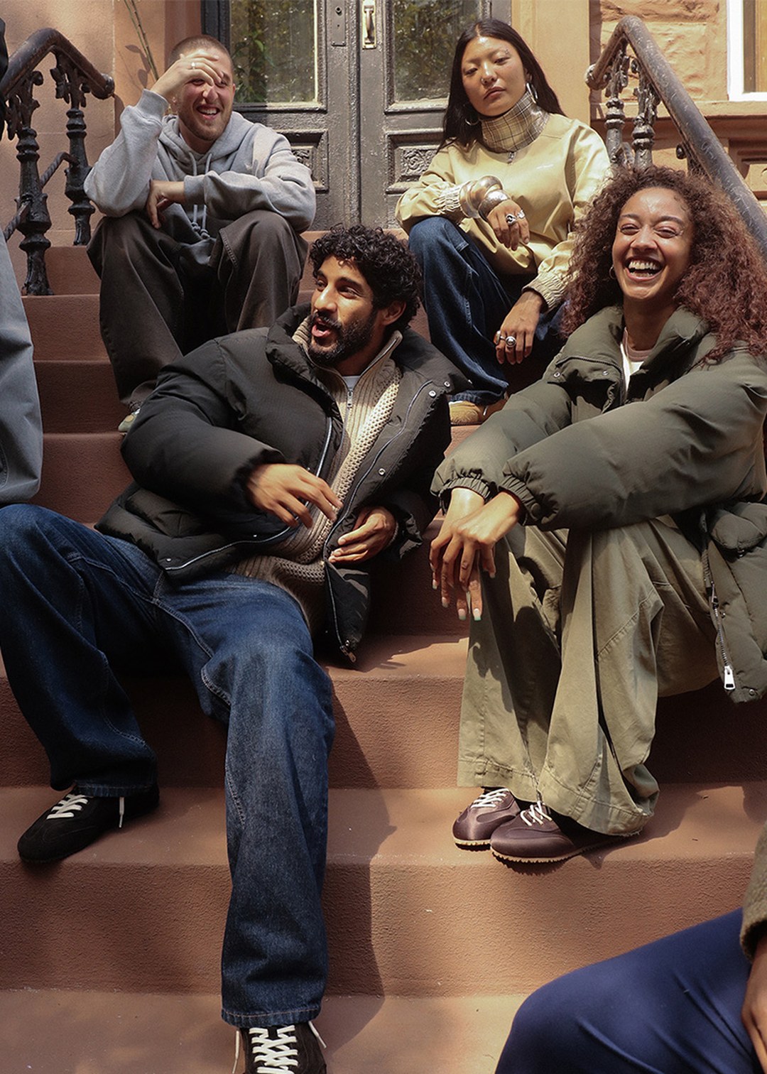 Four people sitting on outdoor steps, laughing and chatting. They are dressed in casual, warm clothing on a sunny day.