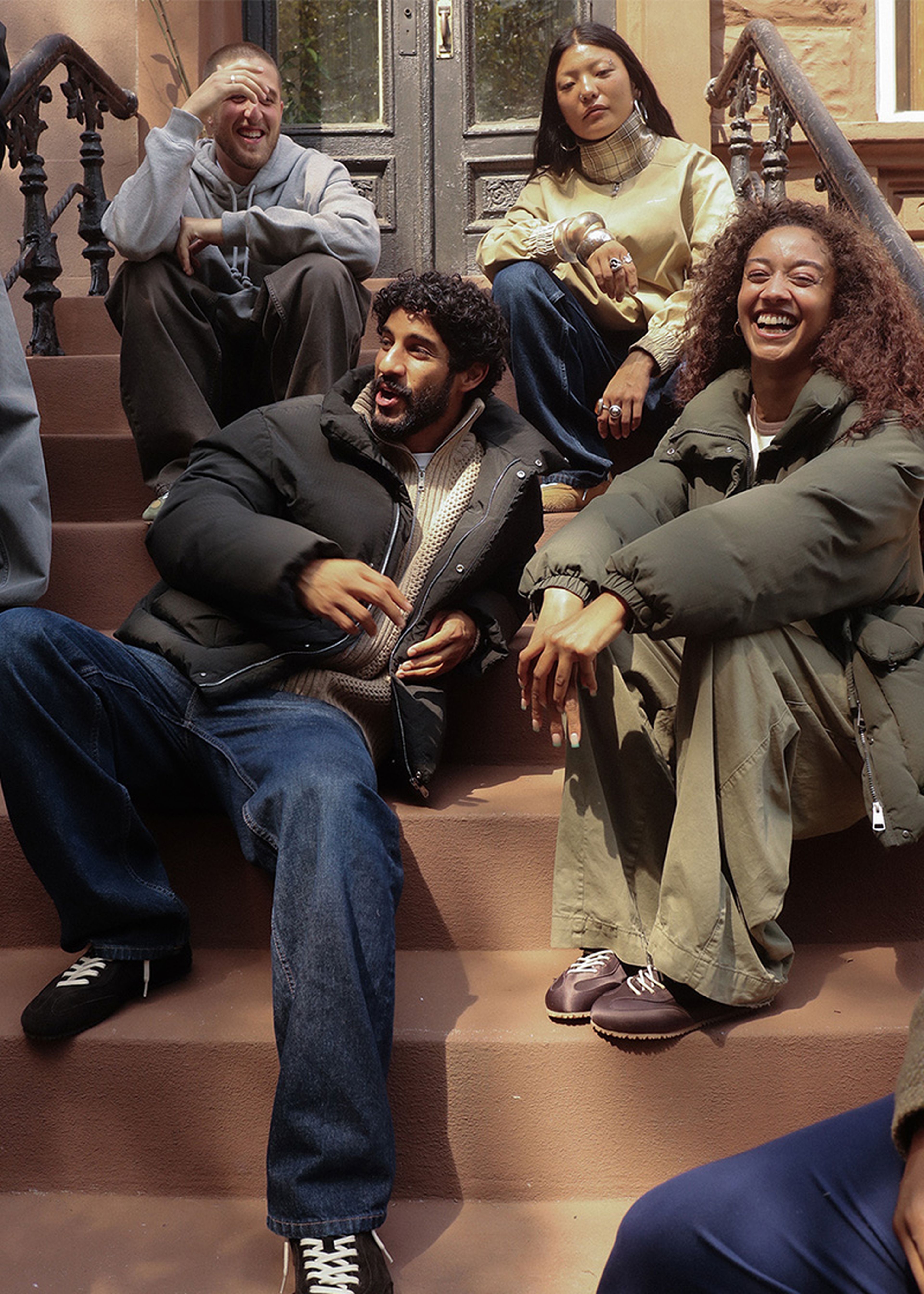 Four people sitting on outdoor steps, laughing and chatting. They are dressed in casual, warm clothing on a sunny day.