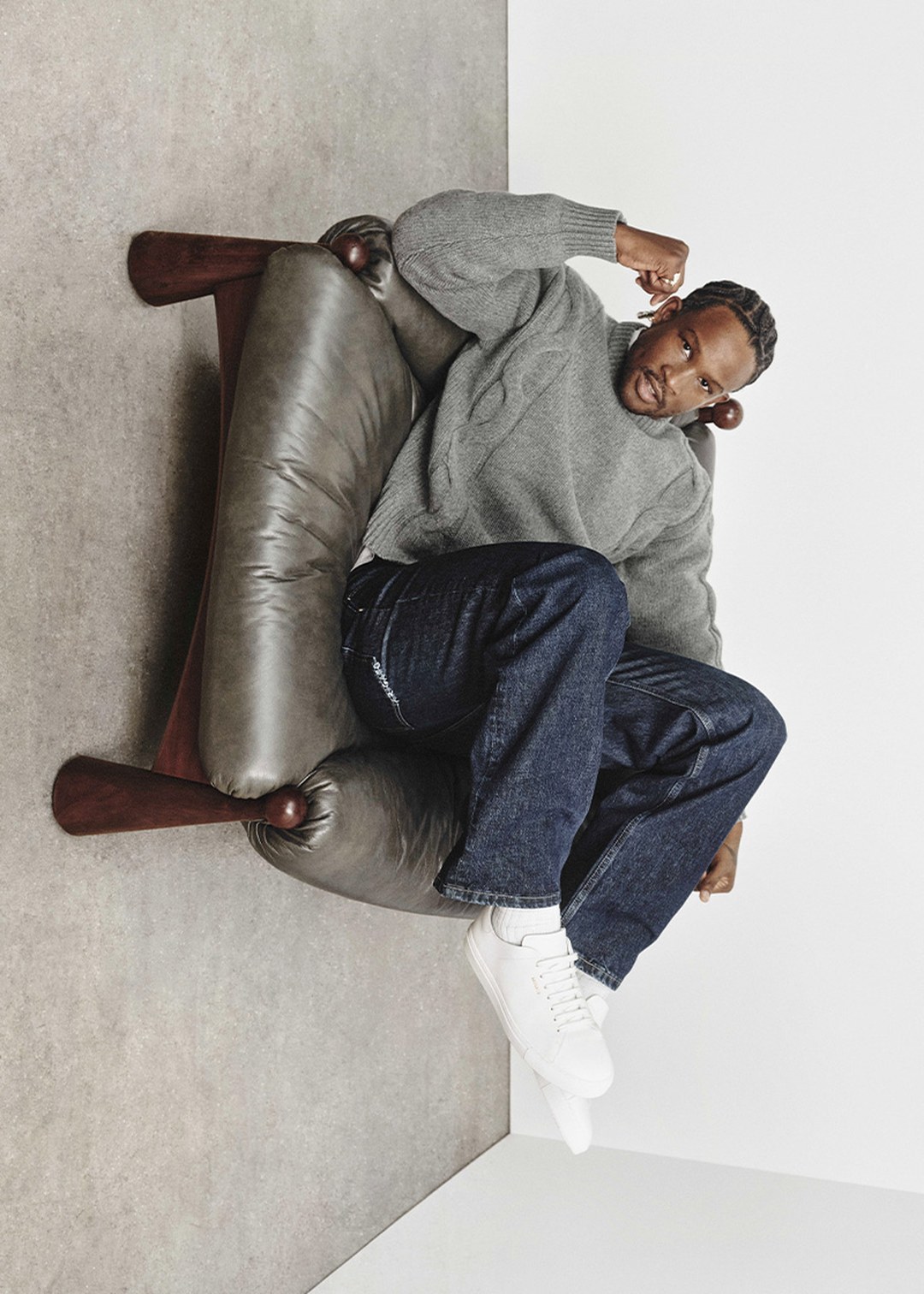 Man in a gray sweater and jeans sits on a leather chair with one arm raised. He's wearing white sneakers, positioned against a minimalist backdrop.