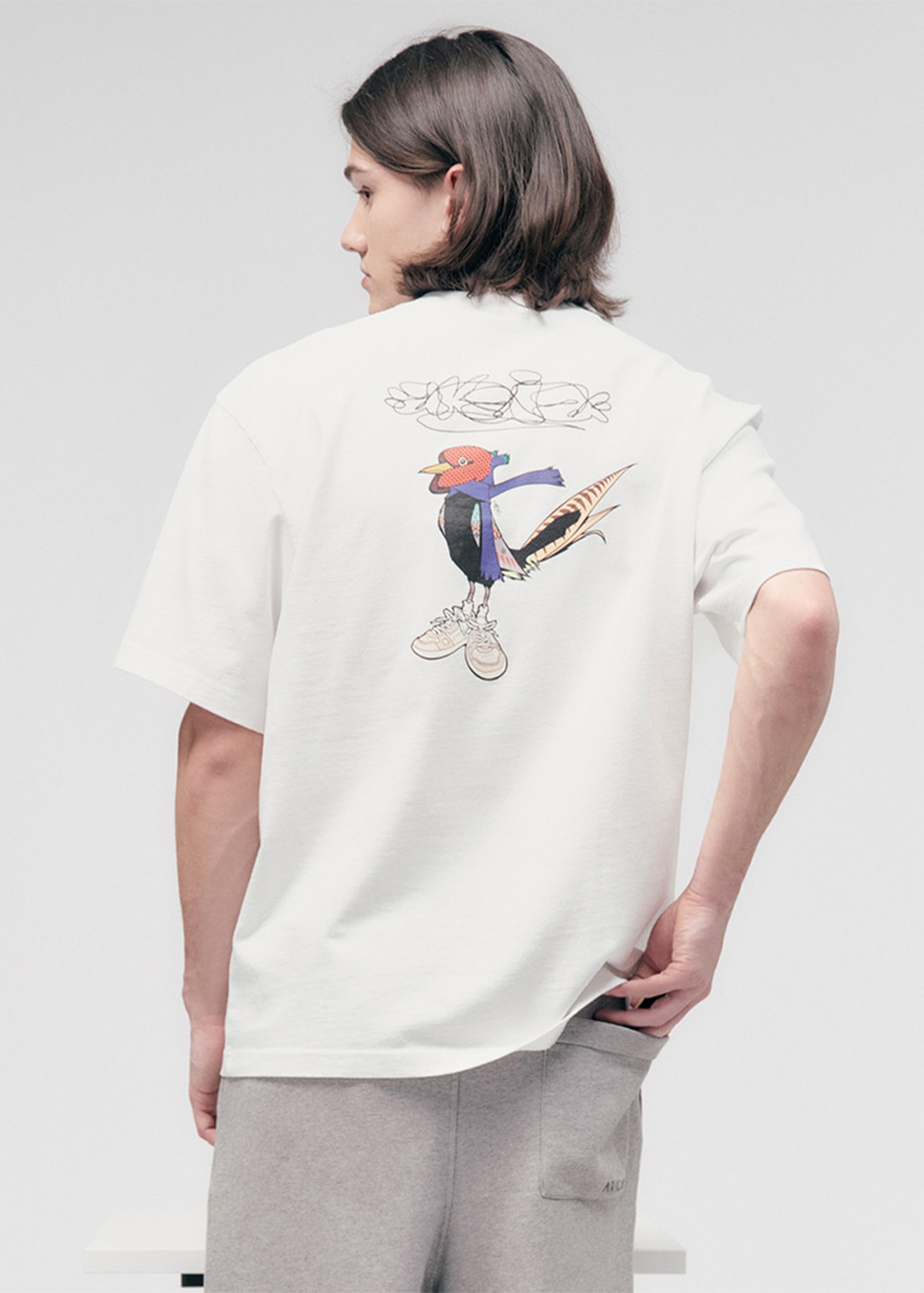 Person with shoulder-length hair wearing a white T-shirt featuring a colorful cartoon bird design on the back, and gray pants.