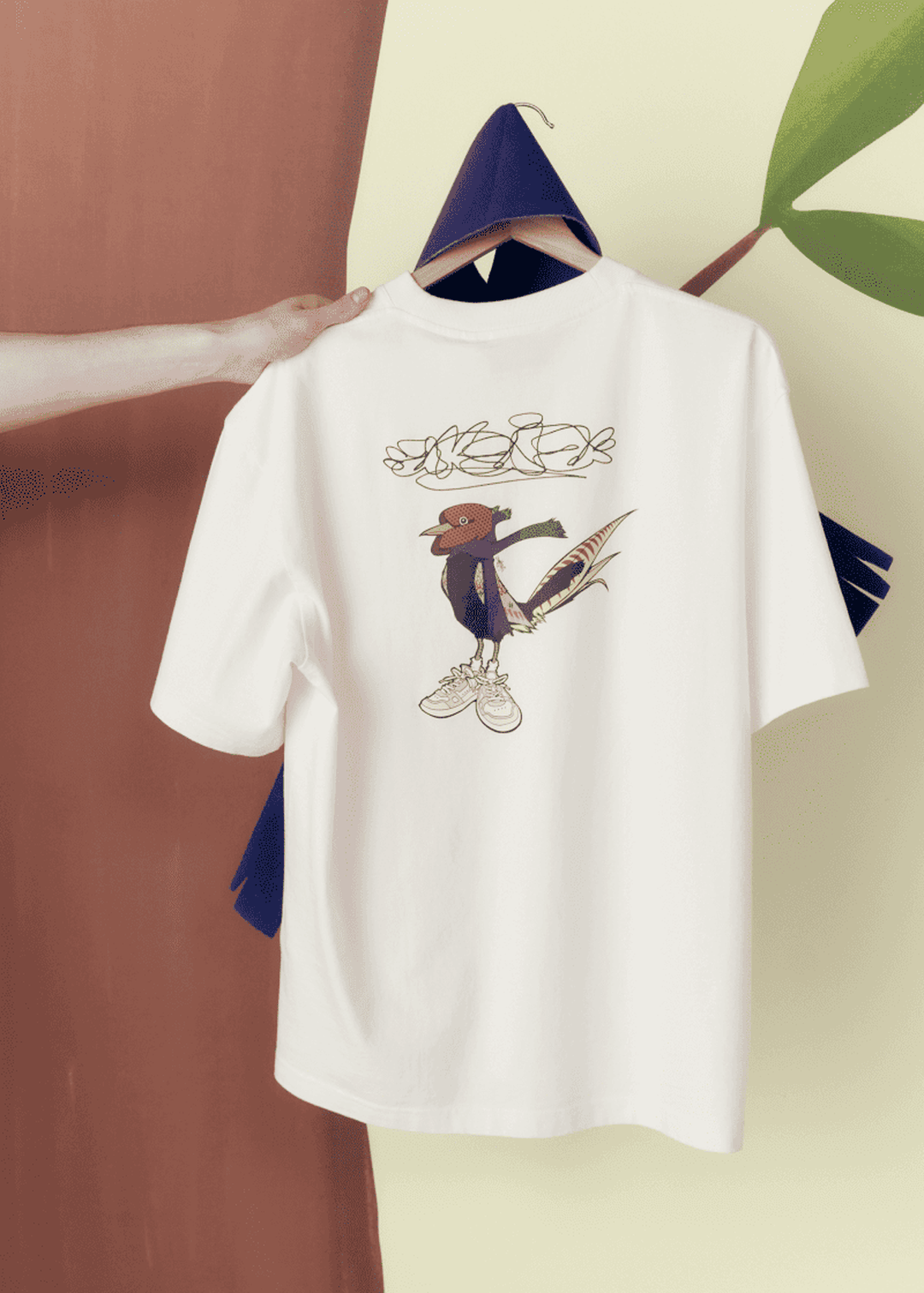 A hand holds a white t-shirt with a bird design wearing sneakers, hanging on a hook against a two-tone background.