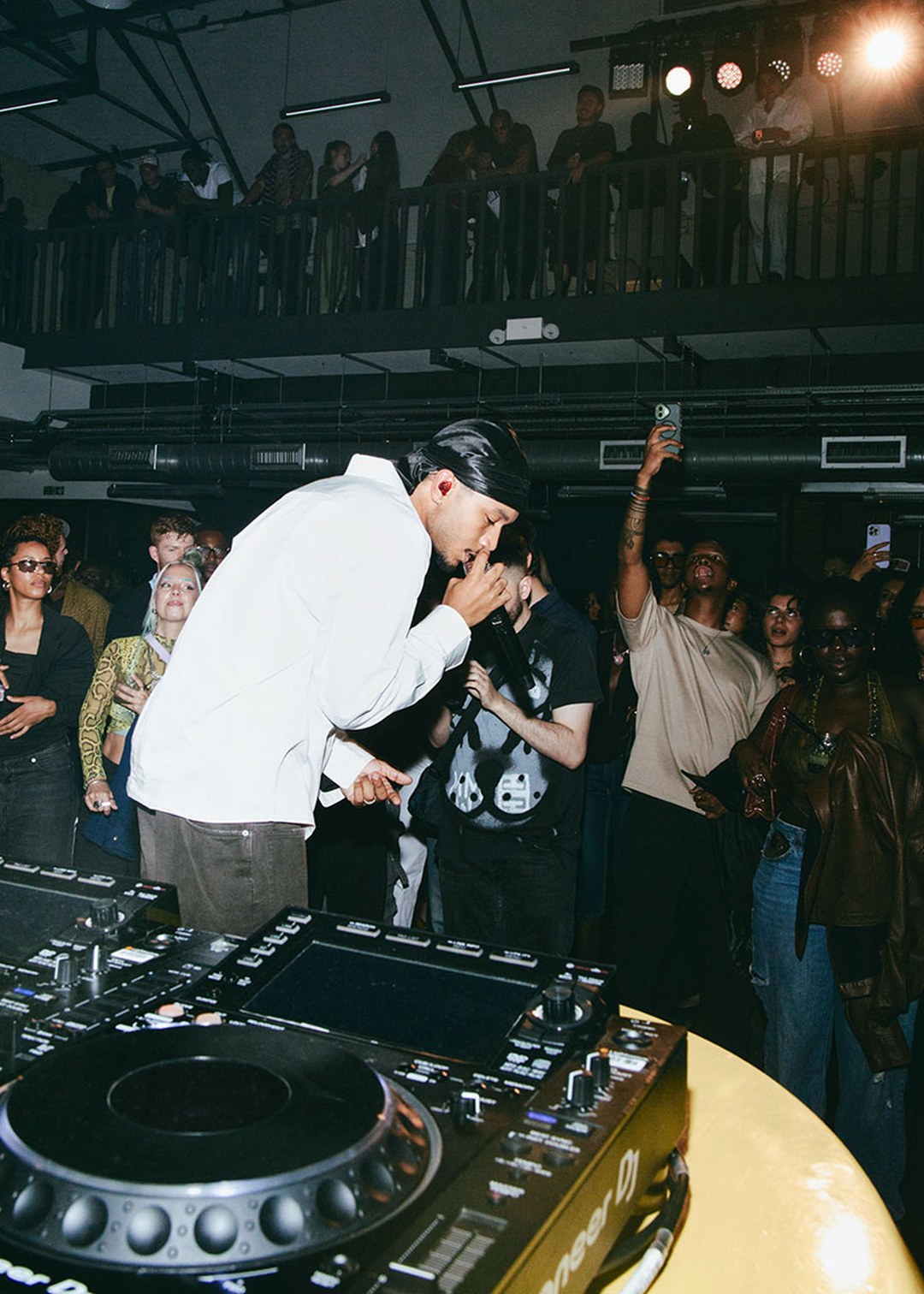 A DJ in a white shirt performs energetically for a lively crowd in a dimly lit club, with people capturing the moment on their phones.