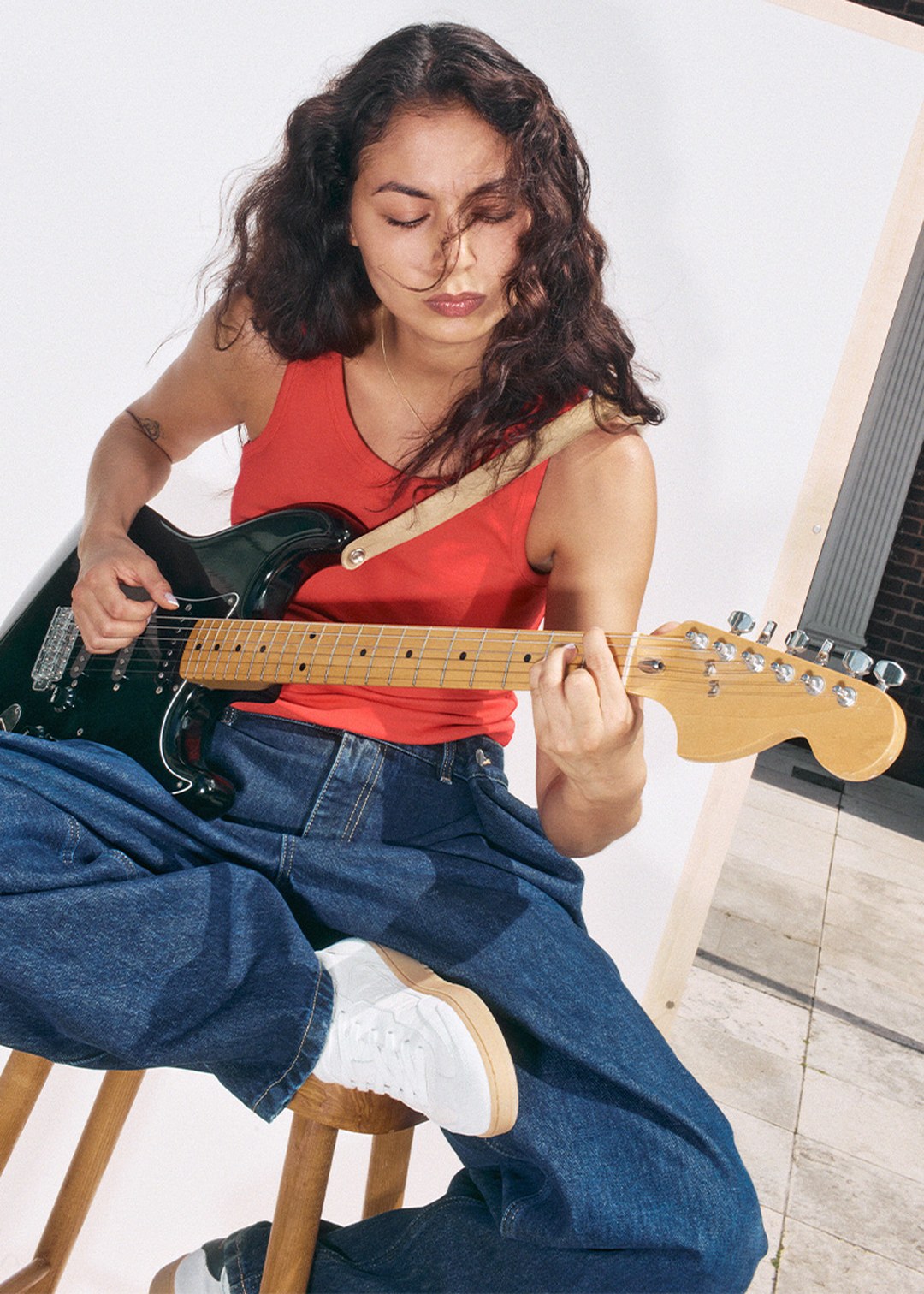 Woman with long curly hair in a red tank top playing an electric guitar while sitting on a stool, against a bright background.
