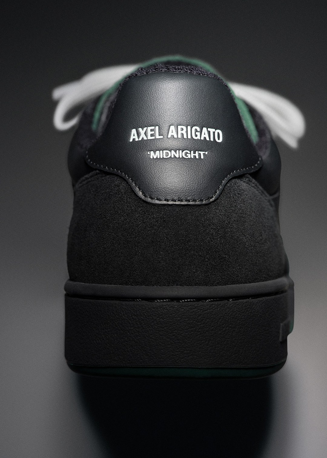 Close-up of the back of a black Axel Arigato sneaker with "MIDNIGHT" written on the heel tab, featuring white laces in the background.