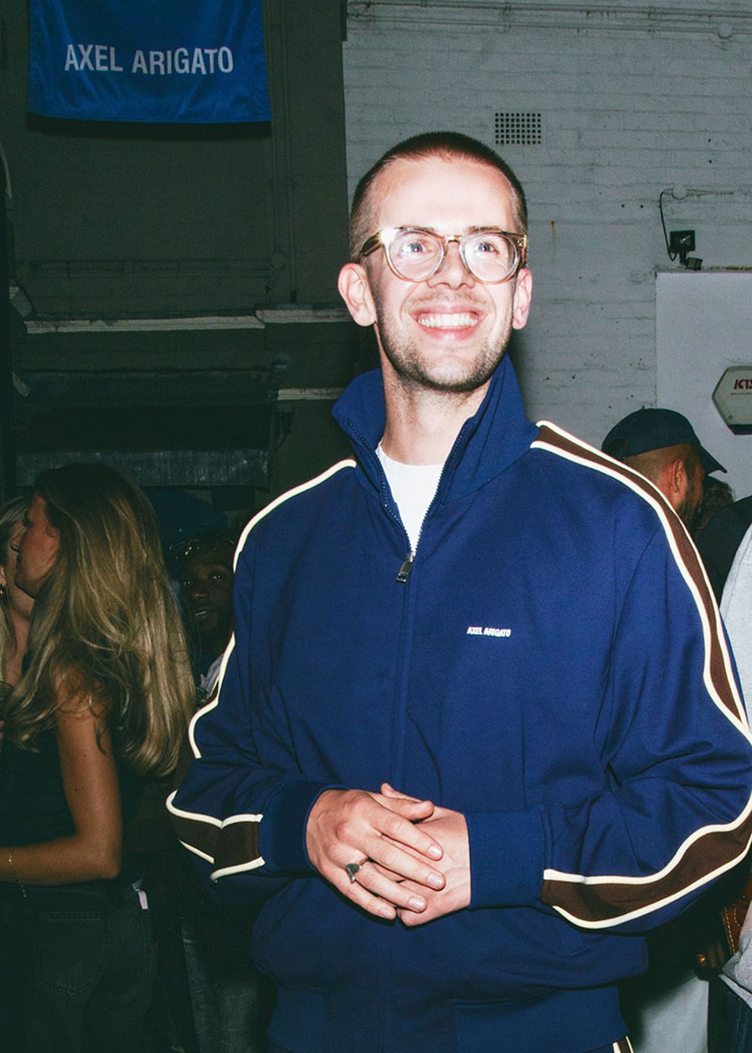 A person wearing glasses and a blue jacket smiles at an event. A sign above reads "AXEL ARIGATO." People are gathered in the background.