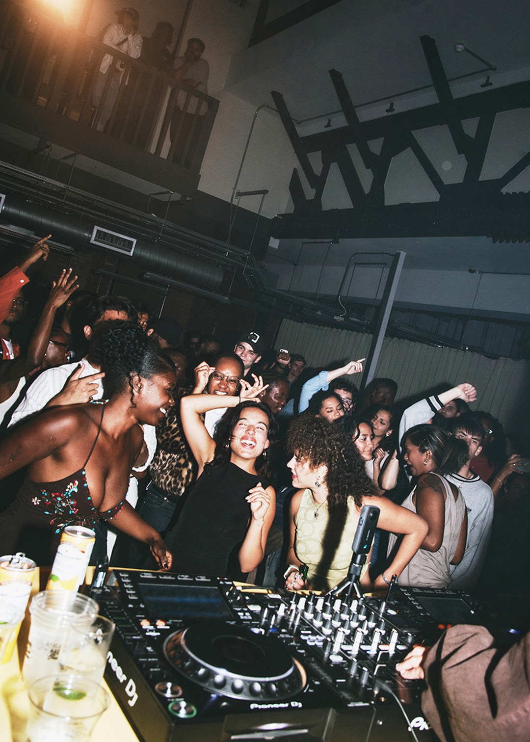 A lively crowd dances and enjoys music in a dimly lit club, with a DJ booth in the foreground and people smiling and raising their hands.
