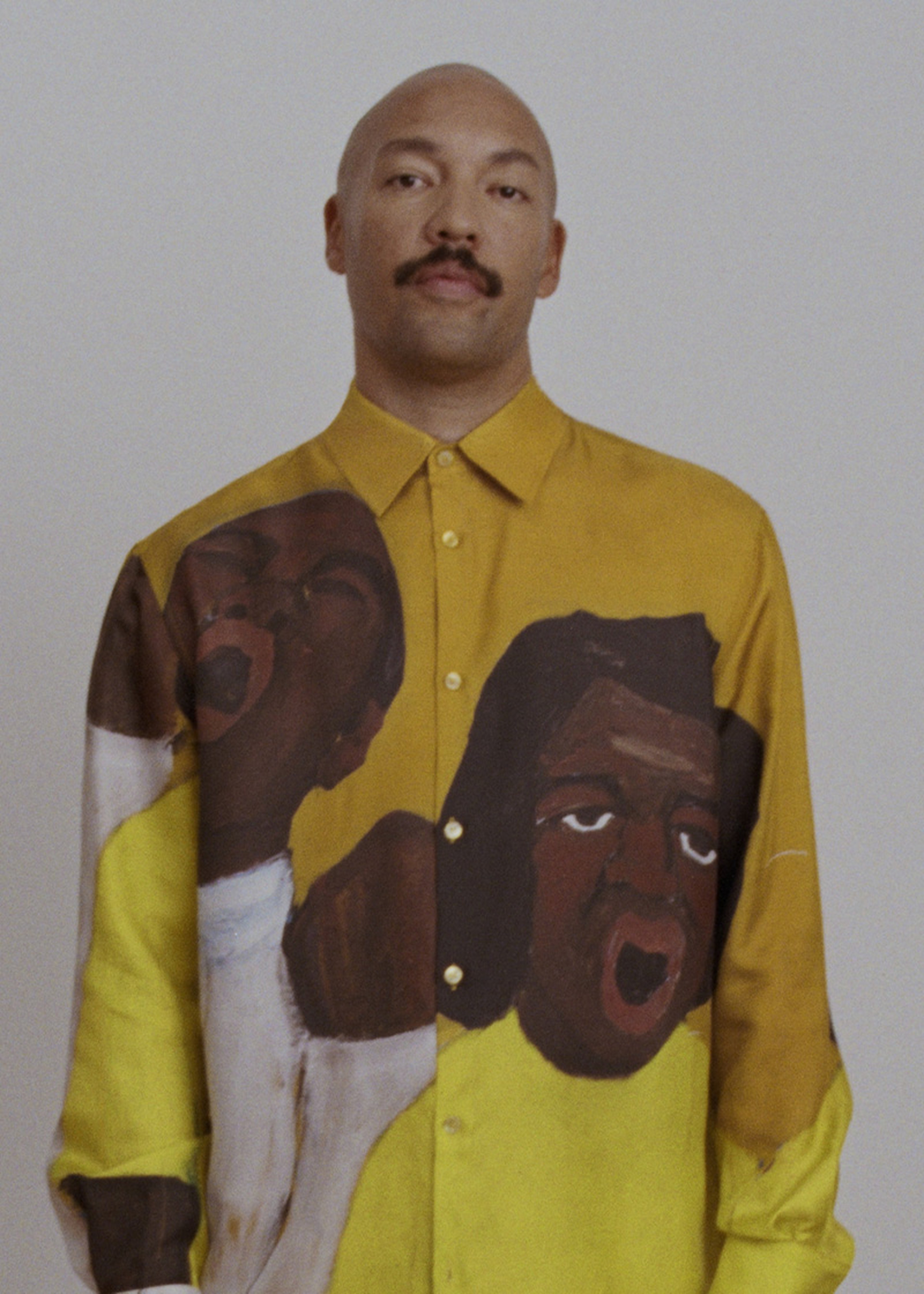 A person wearing a yellow shirt with abstract faces printed on it, standing against a plain background.
