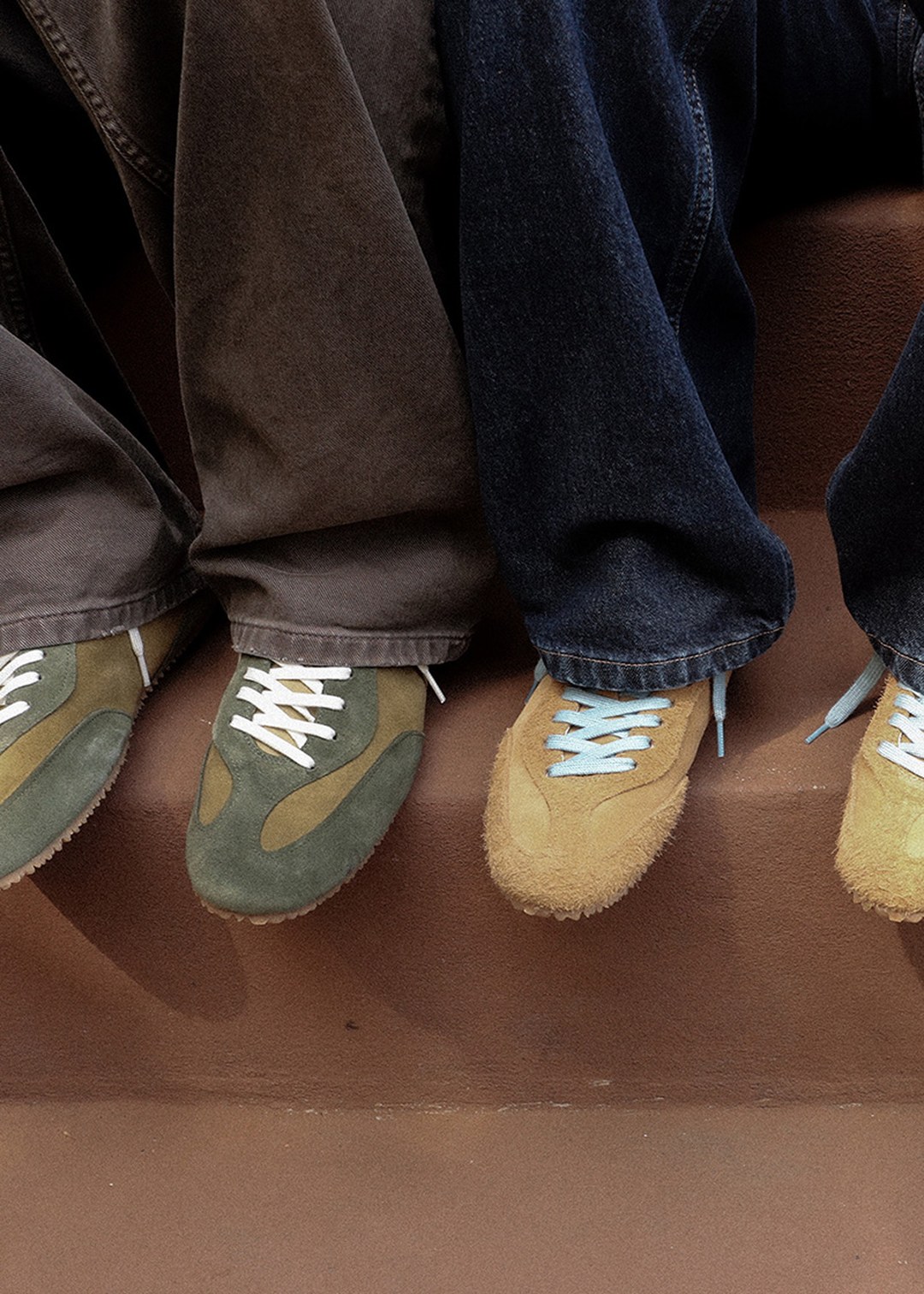 Three pairs of feet in brown suede sneakers and jeans sit on a step, showcasing casual footwear and clothing styles.