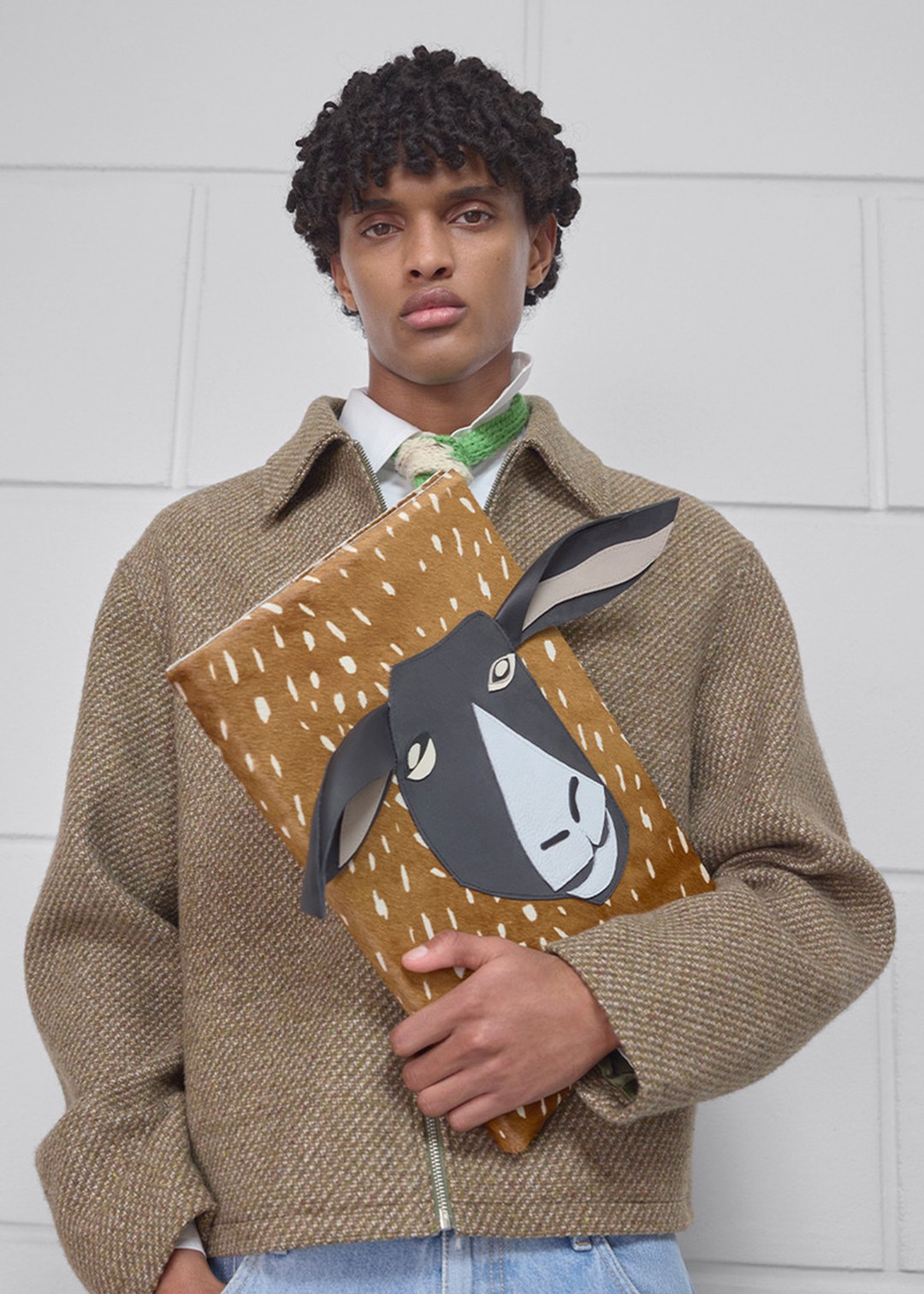 A person in a brown jacket holds a tan clutch with a black and white animal face design, standing against a light wall.