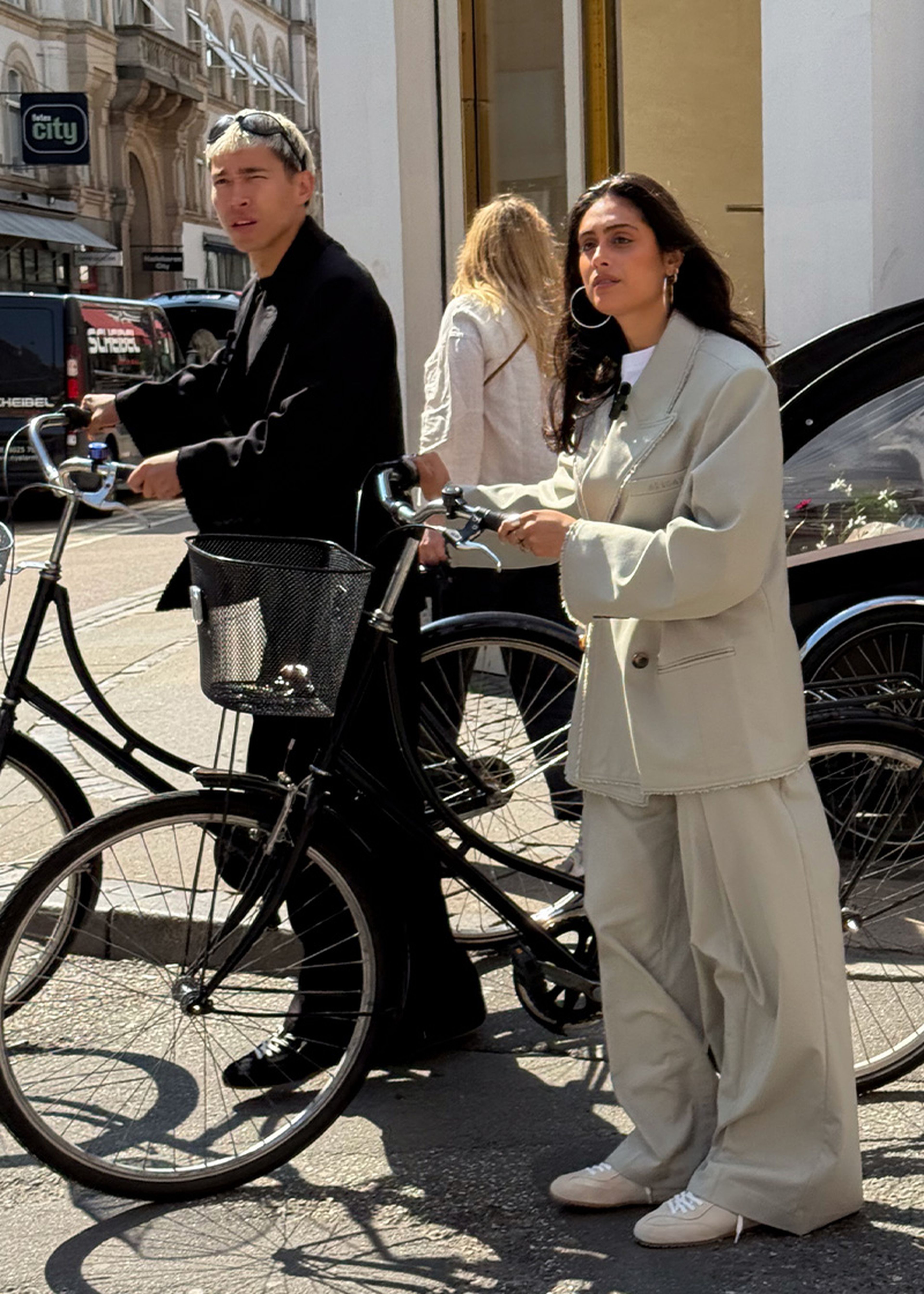 Two people with bicycles stand on a city street. The woman wears an oversized suit, and the man is in black, both looking to the side.