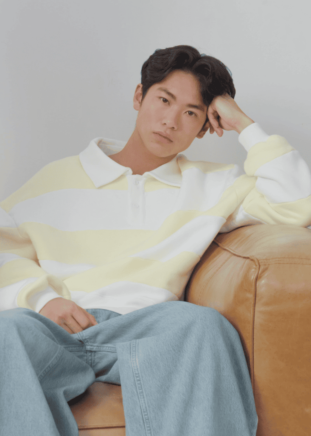 Person in a yellow and white striped sweater and jeans sits on a brown sofa, resting their head on their hand, with a neutral expression.