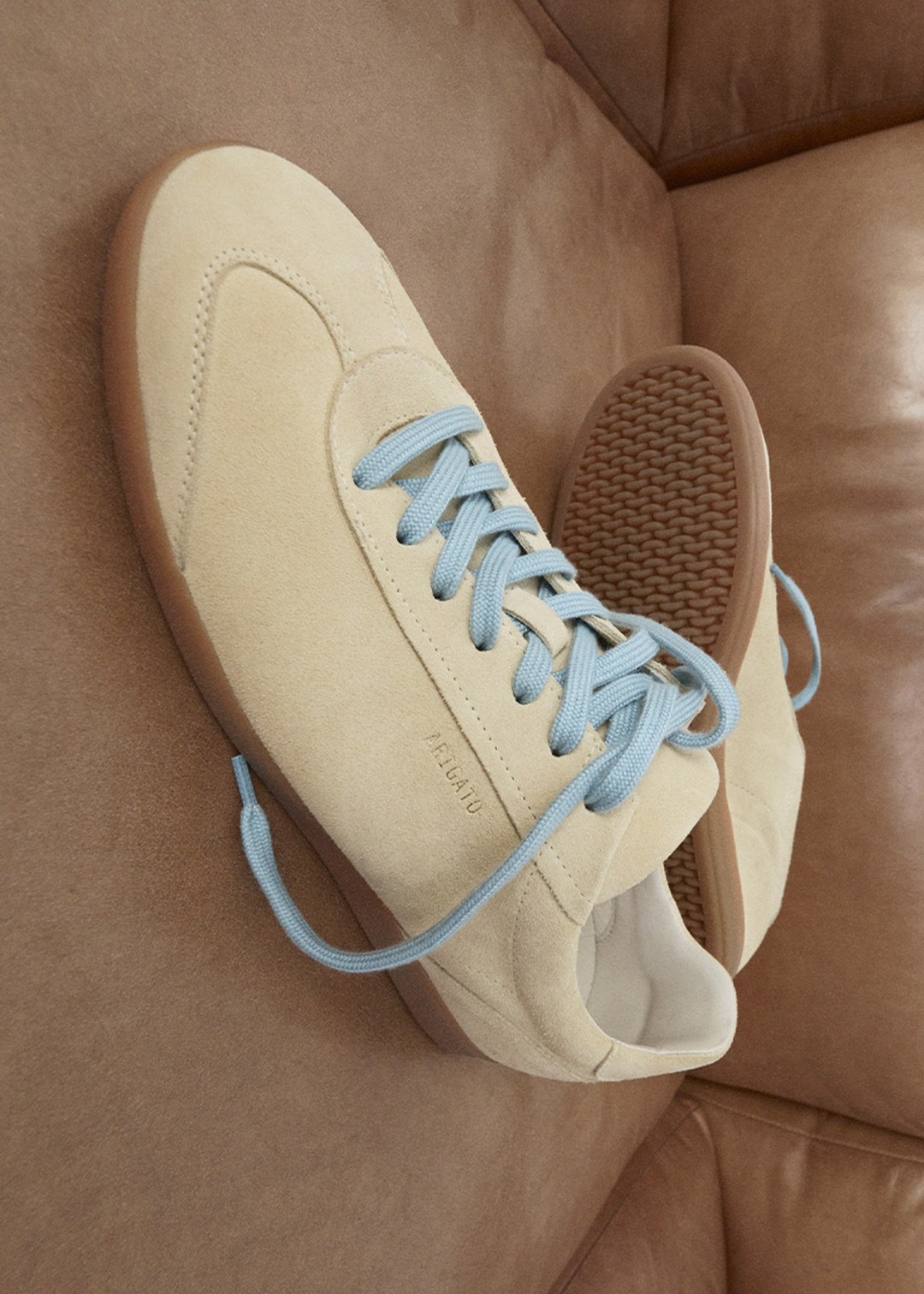 Beige suede sneakers with light blue laces and gum soles, placed on a brown leather surface.
