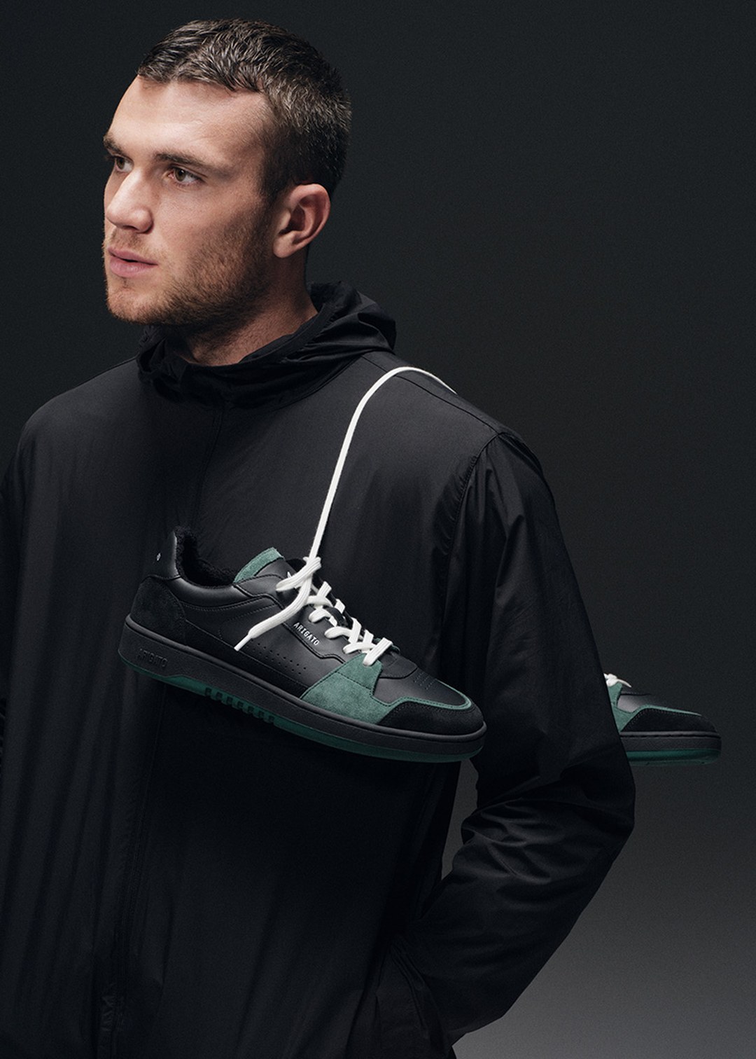 A man in a black hoodie poses with sneakers slung over his shoulder against a dark background.