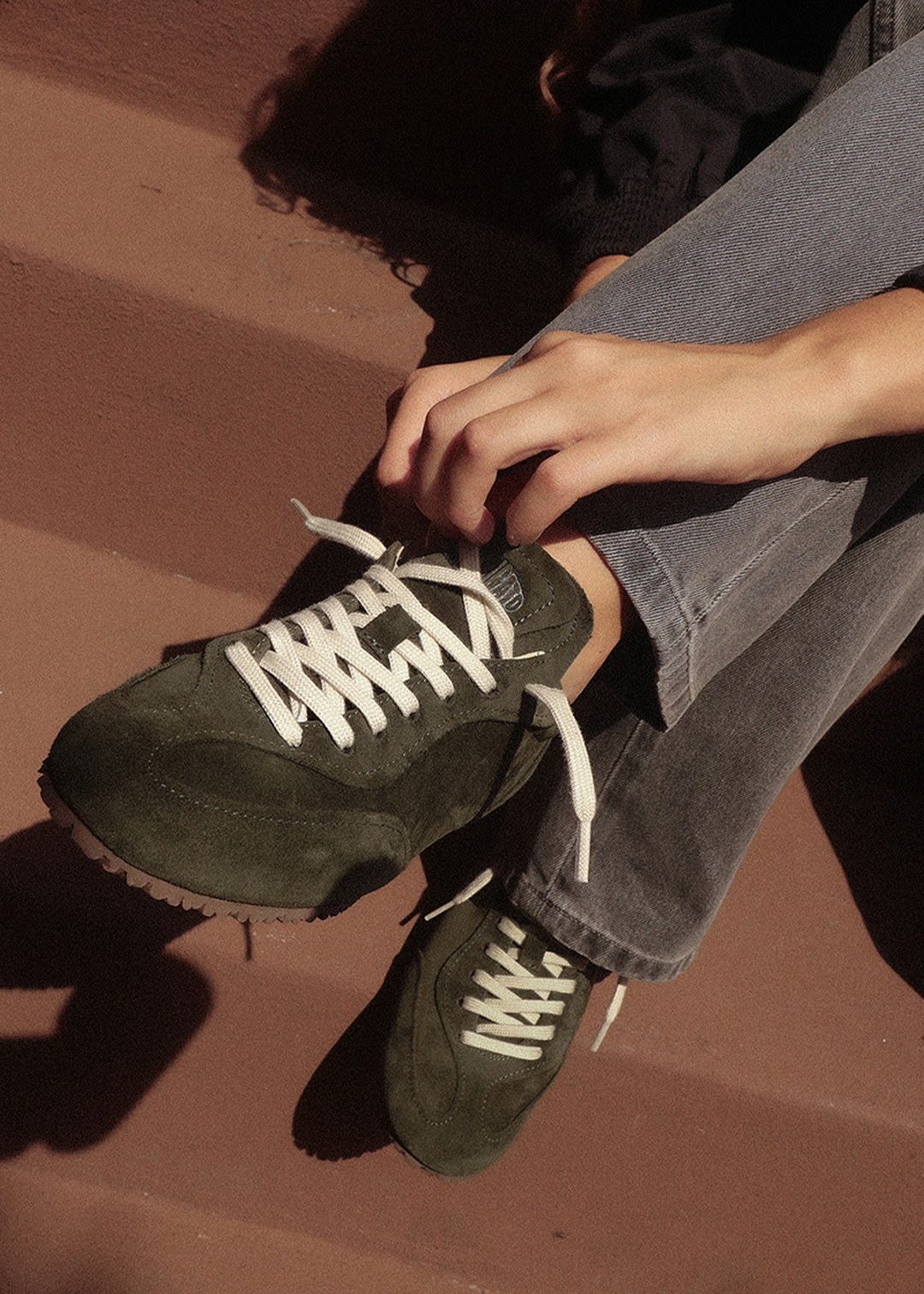 Person sitting on stairs, adjusting a dark green sneaker with white laces, wearing blue jeans.