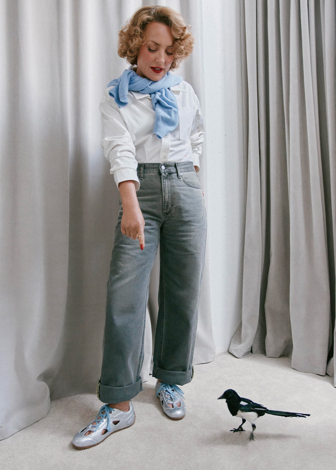 Woman in white shirt and gray jeans points at a bird on the floor. She wears silver shoes and a blue scarf, standing in a room with gray curtains.