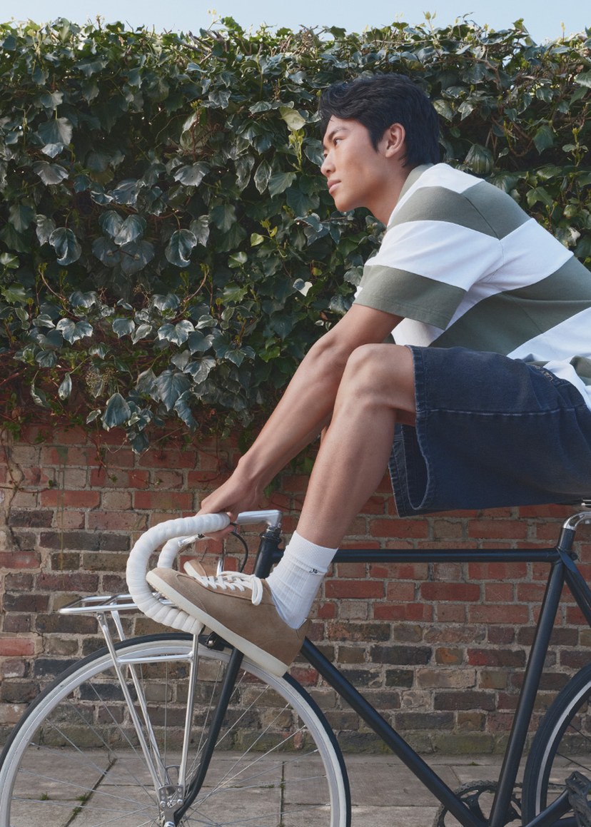 Person in a striped shirt and denim shorts balancing on a stationary bicycle, with greenery and a brick wall in the background.