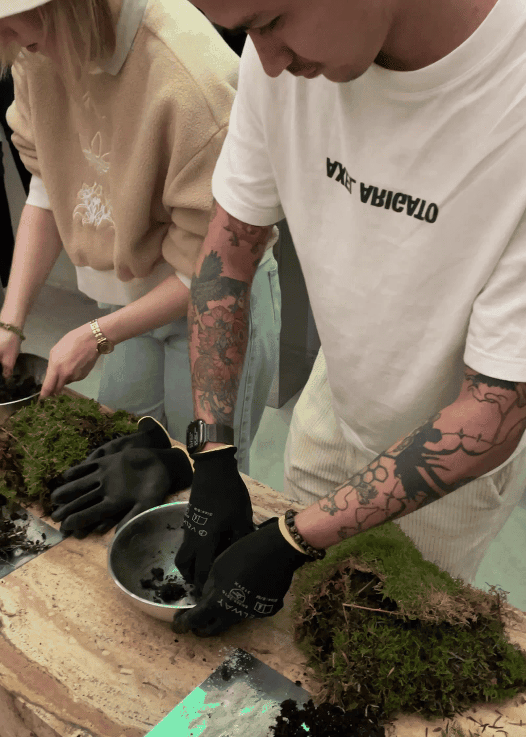 People with tattoos crafting small gardens in trays, wearing gloves, surrounded by soil and moss on a table.