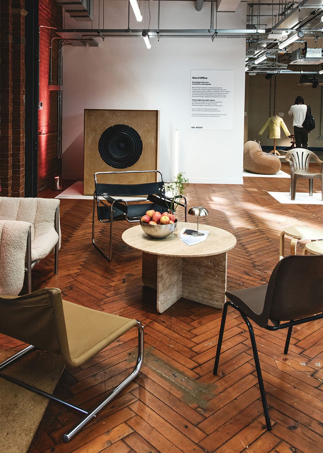 Modern living room with stylish chairs, a round coffee table with a fruit bowl, and a large speaker. Exposed brick wall and wooden floor.