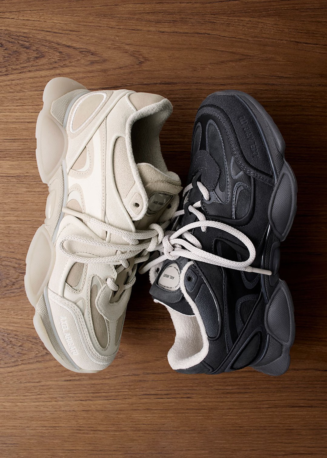 A pair of sneakers on a wooden floor; one is cream-colored, and the other is dark gray, both with chunky soles and intricate designs.