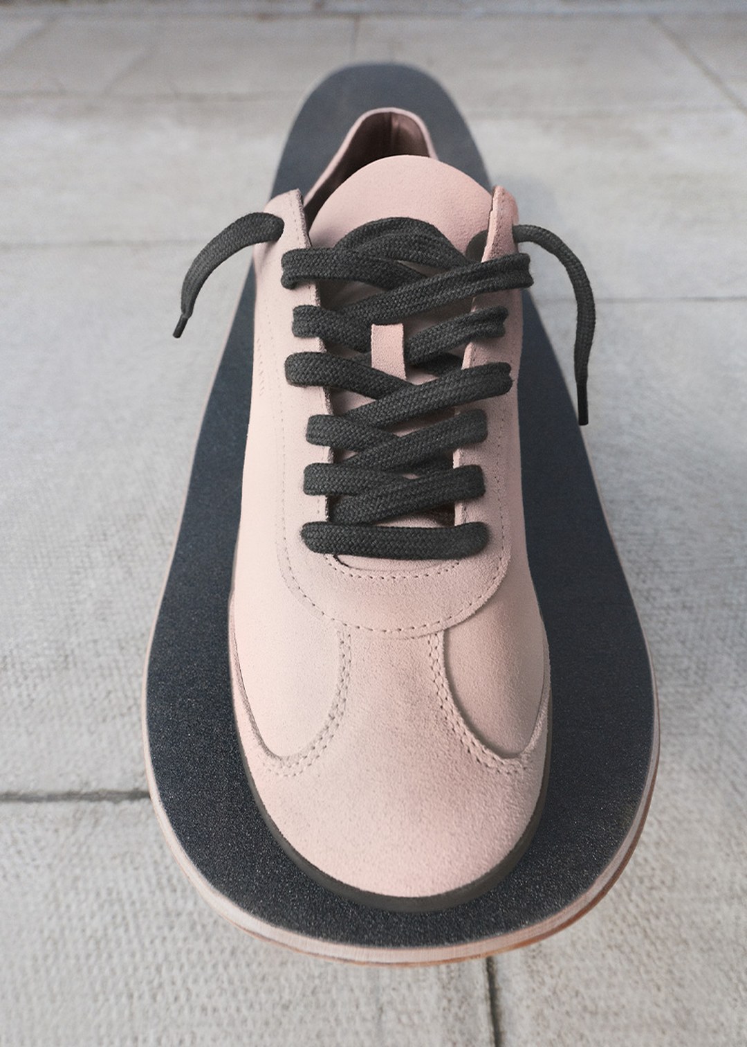 A beige suede sneaker with black laces rests on a skateboard, set against a concrete background.