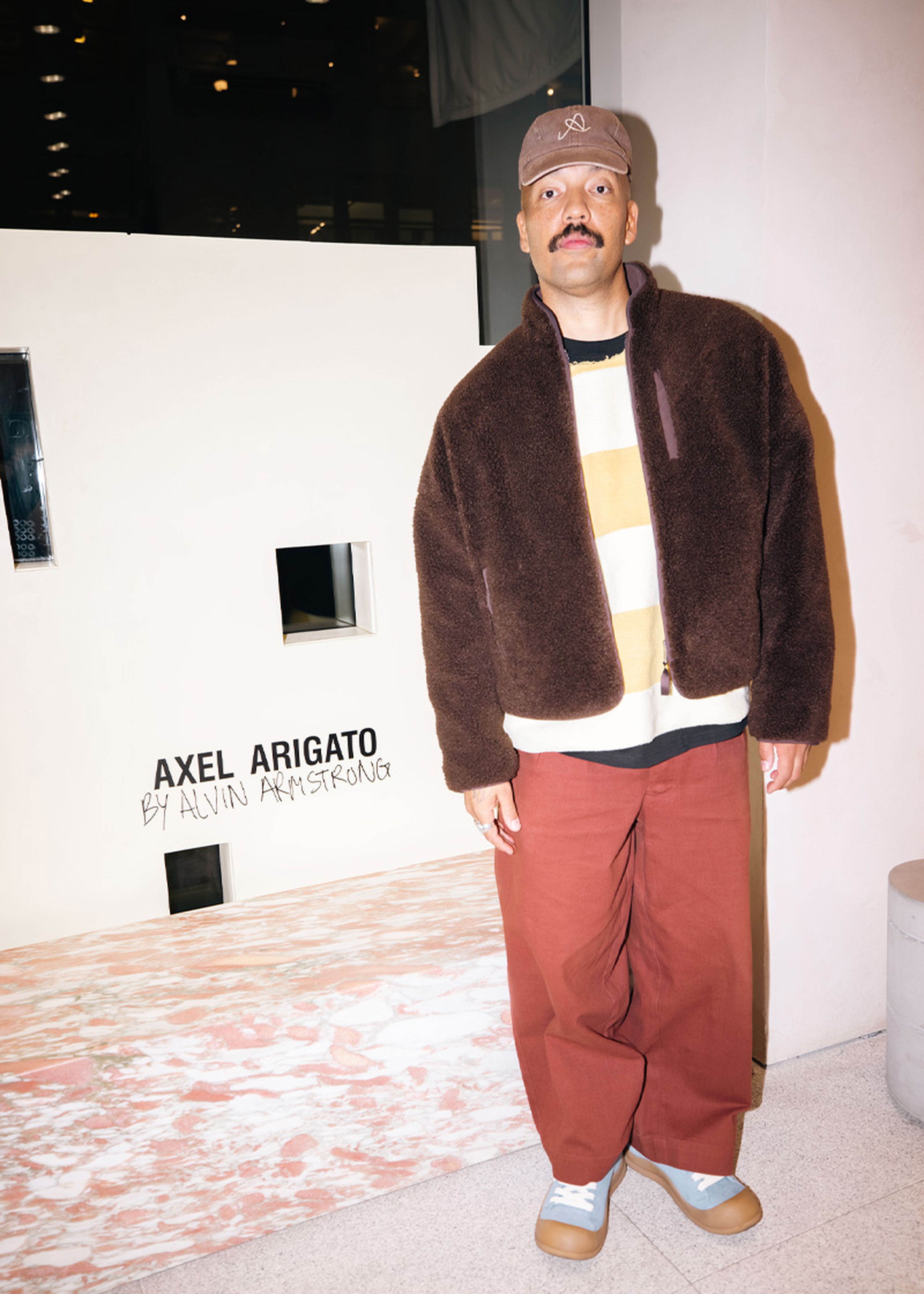 Man in a brown jacket and red pants stands indoors next to a white structure with "AXEL ARIGATO BY ALVIN ARMSTRONG" text.