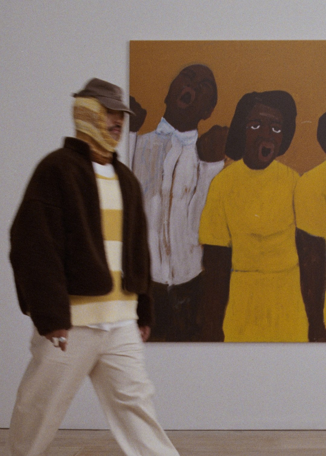 Person in layered clothing walks past a painting of three expressive figures in a gallery setting.