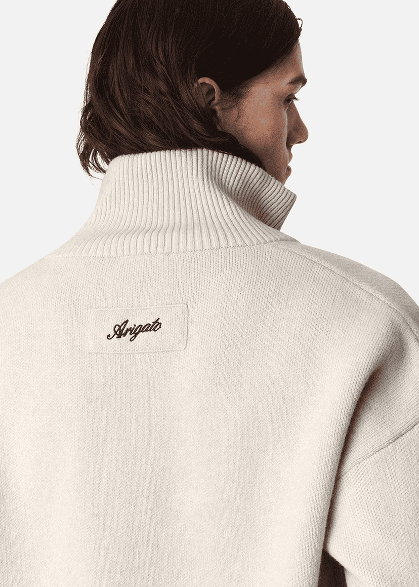 A person with long hair in a cream high-collar sweater, featuring "Arigato" embroidered on the back.