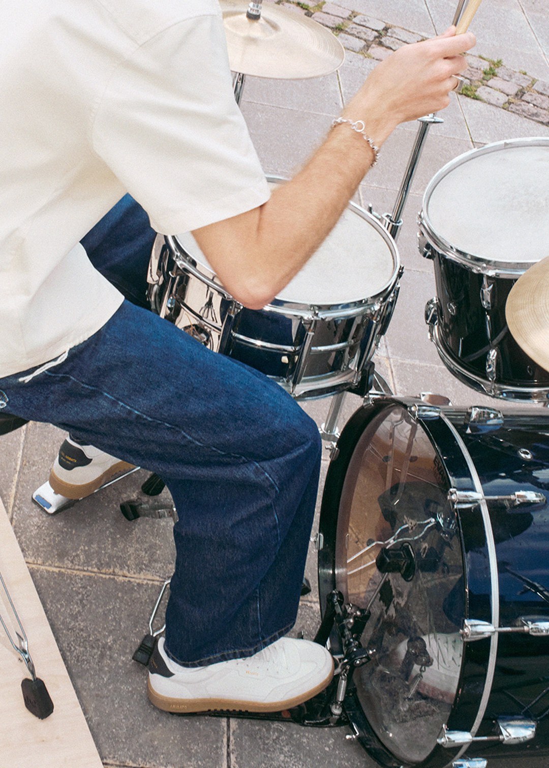 Person playing a drum set outdoors, wearing a white shirt, blue jeans, and white sneakers. Drums include a bass drum and snare.