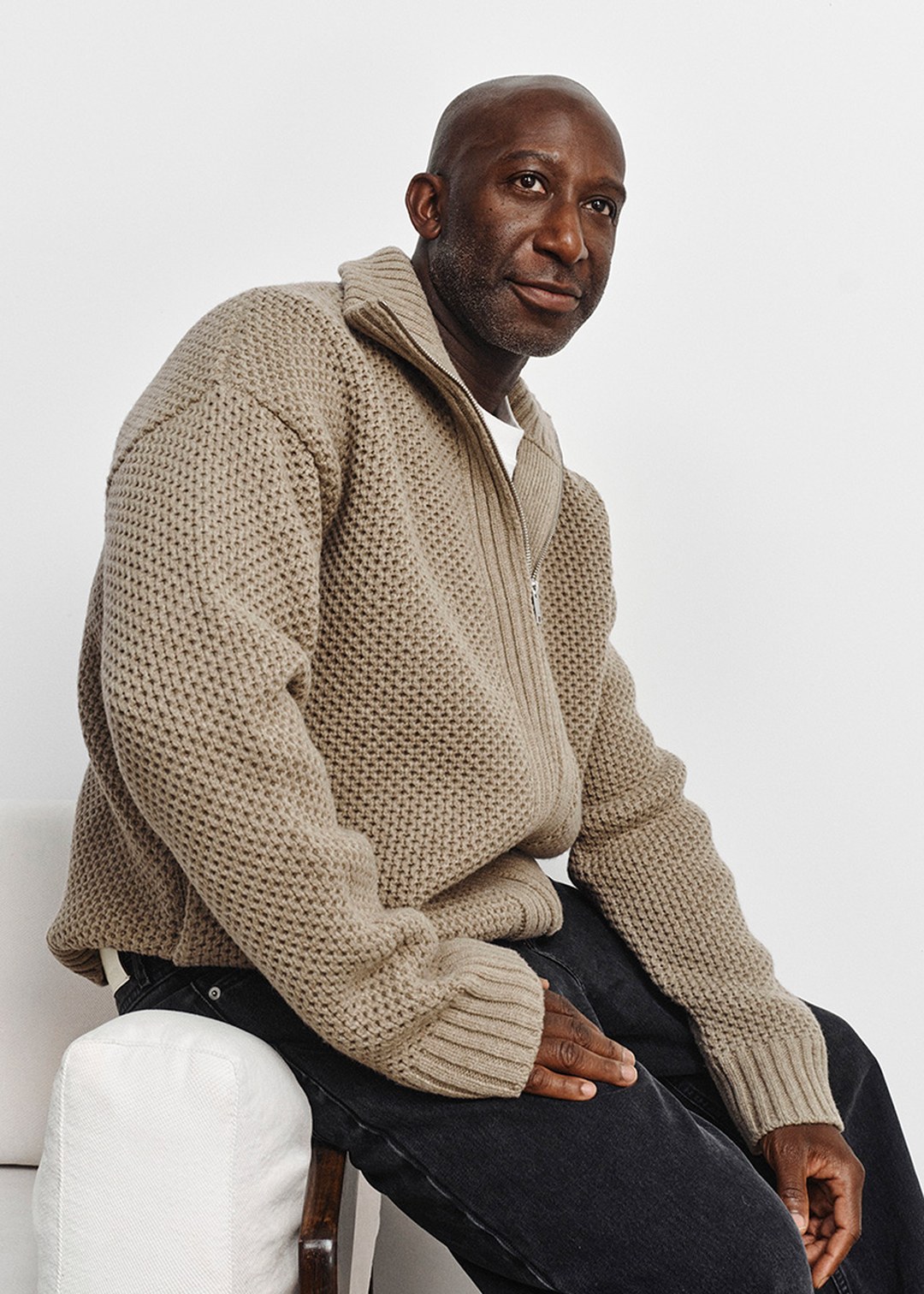 Man seated on a chair, wearing a chunky beige knitted sweater and dark pants, looking to the side. White background.