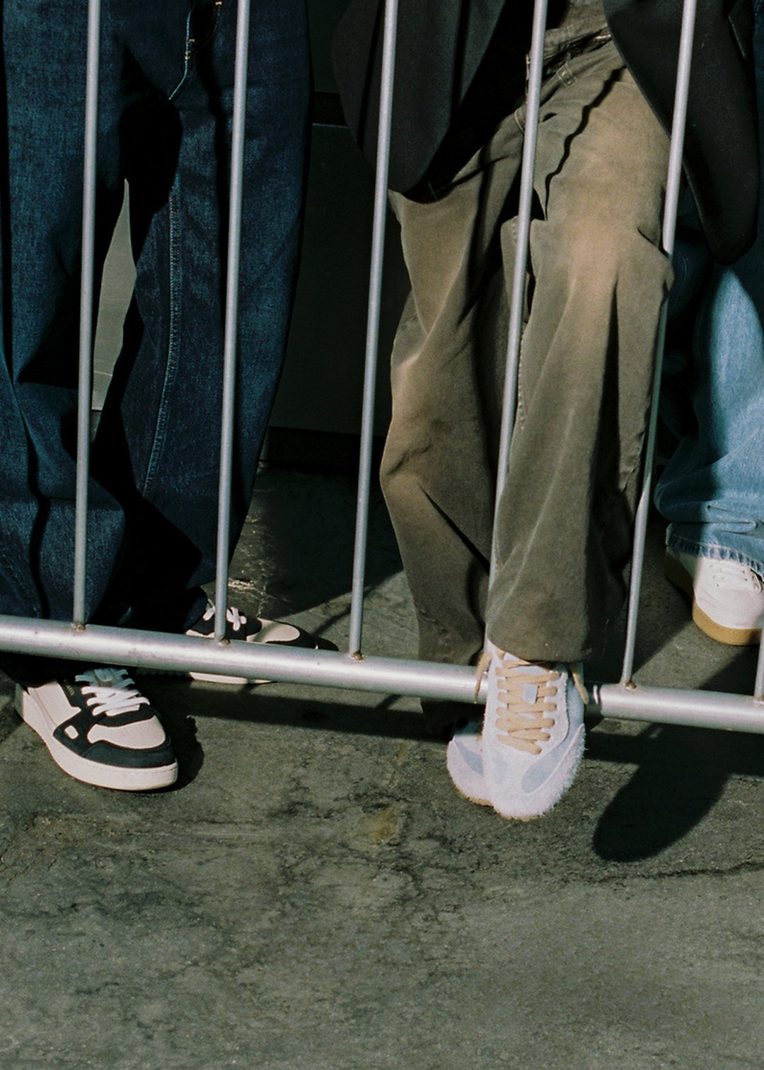 Three people standing behind a metal fence, wearing casual sneakers and pants, on a concrete surface.