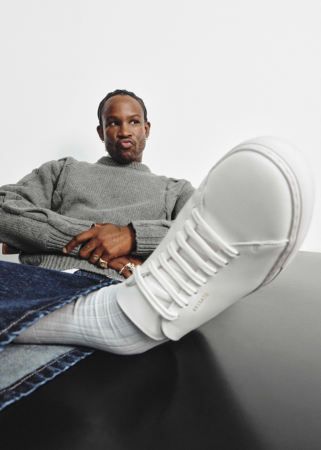 Man in a gray sweater and jeans sits with one leg raised, showcasing a white sneaker. The background is white, giving a minimalist look.